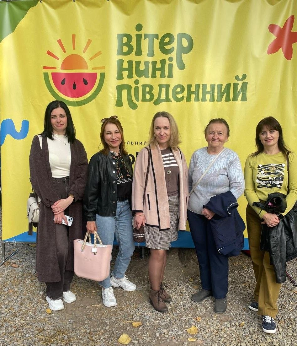 П'ять усміхнених жінок стоять у ряд на фоні великого жовтого банера з логотипом фестивалю «вітер нині південний». На банері зображено сонце, що сходить над червоною скибкою херсонського кавуна. Жінки одягнені в сучасний повсякденний одяг і тримають сумочки