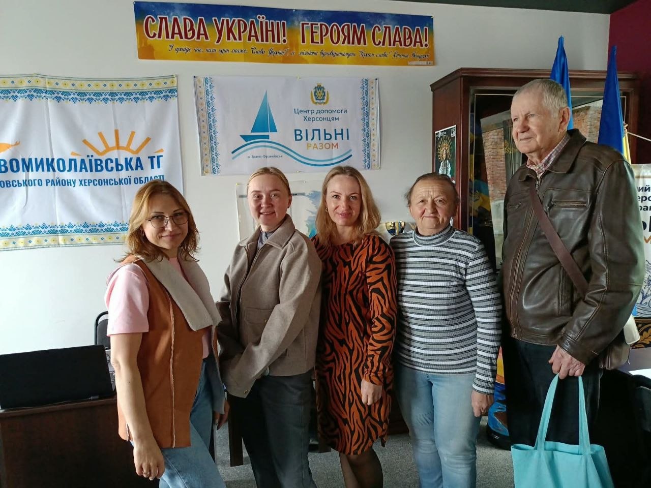 Група з п'яти людей — чотири жінки та літній чоловік у шкіряній куртці зі світло-блакитною торбинкою в руках — позують у приміщенні хабу для переселенців. Вони стоять у ряду перед камерою. На стіні позаду закріплено плакат Центру допомоги херсонцям у м. Івано-Франківськ «ВІЛЬНІ РАЗОМ», символіку Новомиколаївської ТГ Скадовського району, гасло «СЛАВА УКРАЇНІ! ГЕРОЯМ СЛАВА!» та розгорнуто сині прапори.