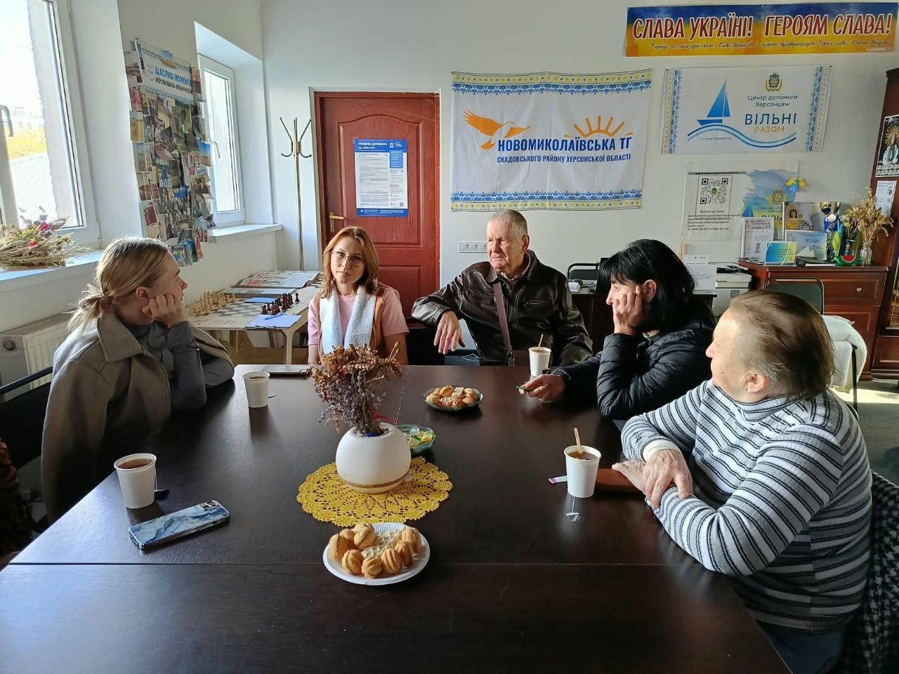 Зображення 1 (5321338921130074106.jpeg) Шестеро людей сидять за круглим темним столом у приміщенні центру допомоги під час спілкування та чаювання. На столі стоять паперові стаканчики з гарячими напоями, тарілки з домашнім печивом у формі горішків, вазочка з сухоцвітами на жовтій мереживній серветці та лежить смартфон. На стінах позаду розміщено патріотичний банер «СЛАВА УКРАЇНІ! ГЕРОЯМ СЛАВА!», символіку Новомиколаївської територіальної громади Скадовського району Херсонської області та прапорець хабу «ВІЛЬНІ РАЗОМ». Біля вікна на додатковому столику видно розкладену шахівницю.