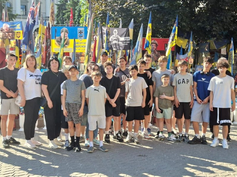 Групове фото великої делегації, що складається переважно з підлітків, дітей та кількох супроводжуючих дорослих (двох жінок та чоловіка у кепці), які вишикувалися в ряд на міській площі чи алеї.  Усі присутні одягнені у літній повсякденний одяг: футболки (зокрема з логотипами GAP, Armani, Puma), шорти та кросівки. Вони дивляться в об'єктив камери. Зйомка ведеться у сонячний день, на бруківці попереду видно довгі тіні.  Безпосередньо за групою облаштовано велике меморіальне місце вшанування пам'яті полеглих захисників України:  Майданчик щільно заповнений десятками синьо-жовтих державних прапорів, а також червоно-чорних та військових стягів на високих флагштоках.  Серед прапорів встановлено великі прямокутні портрети українських військовослужбовців із підписами та біографічними даними. Один із плакатів ліворуч містить жовто-блакитне тло та фото воїна, інший по центру — чорно-біле зображення бійця зі спареним підписом, де вгадується прізвище «Урбан».  Біля підніжжя меморіалу видно квіти та лампадки. На задньому плані височіють крони зелених дерев.