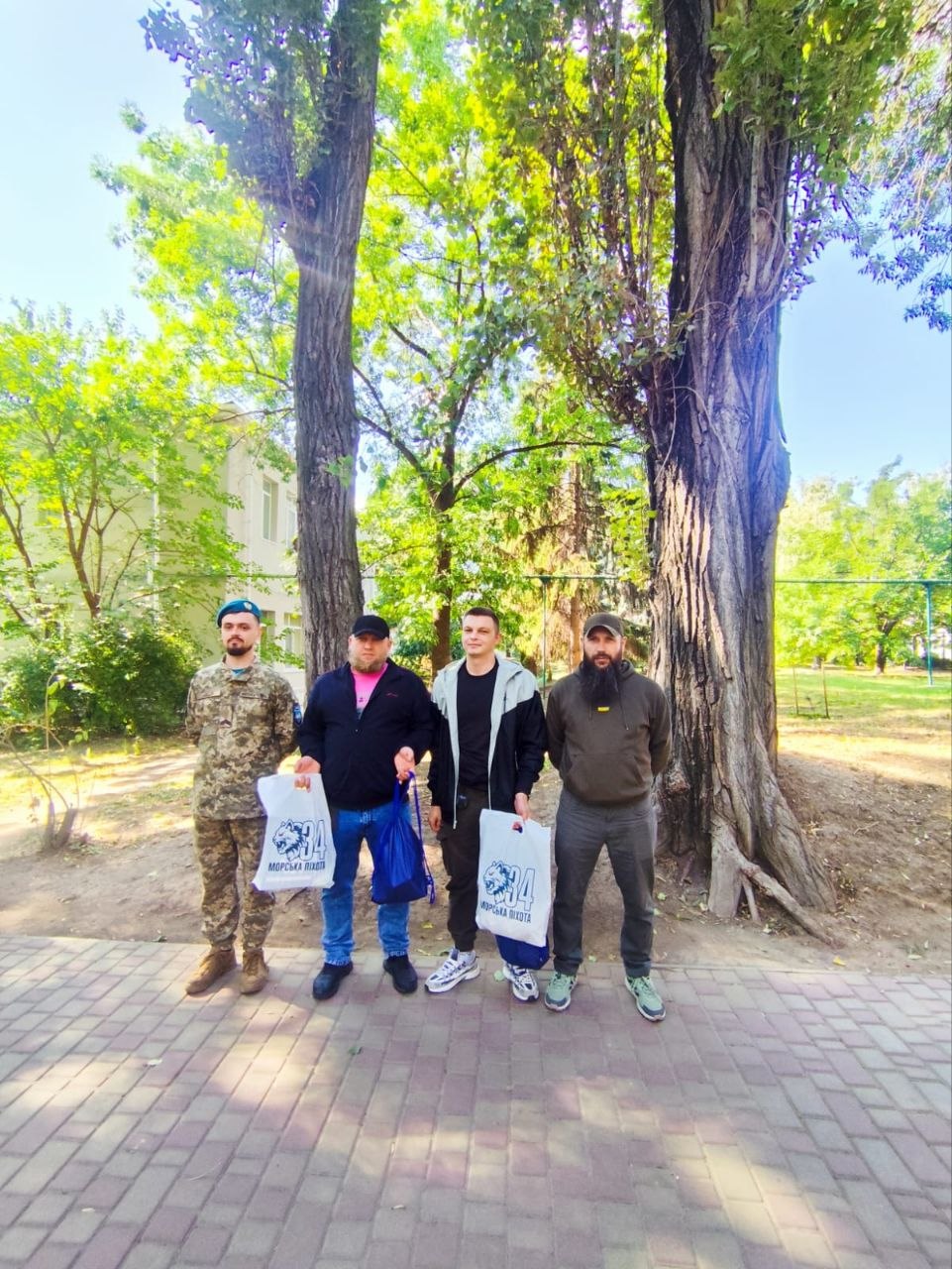 Четверо чоловіків стоять на викладеній бруківкою доріжці в парку між двома високими розлогими деревами. Ліворуч стоїть військовослужбовець у камуфляжній формі та бірюзовому береті морської піхоти. Поруч троє чоловіків у цивільному одязі тримають білі фірмові пакети з емблемою волонтерської або військової підтримки та написом «МОРСЬКА ПІХОТА», а також синю торбинку. На фоні видно зелену паркову зону та будівлю.