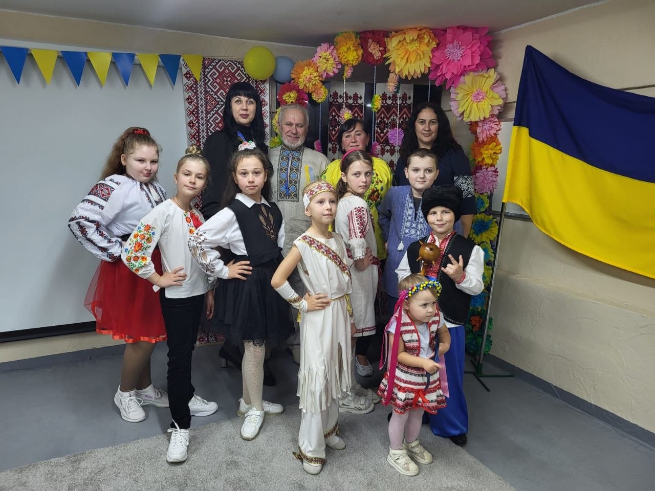 Групове фото юних артистів у сценічних строях разом із наставниками біля фотозони з великими різноколірними паперовими квітами, вишитими рушниками та синьо-жовтим стягом. Діти одягнені у вишиванки, козацькі шаровари, сценічні сукні та стилізоване біле вбрання, а дорослі позаду щиро посміхаються.