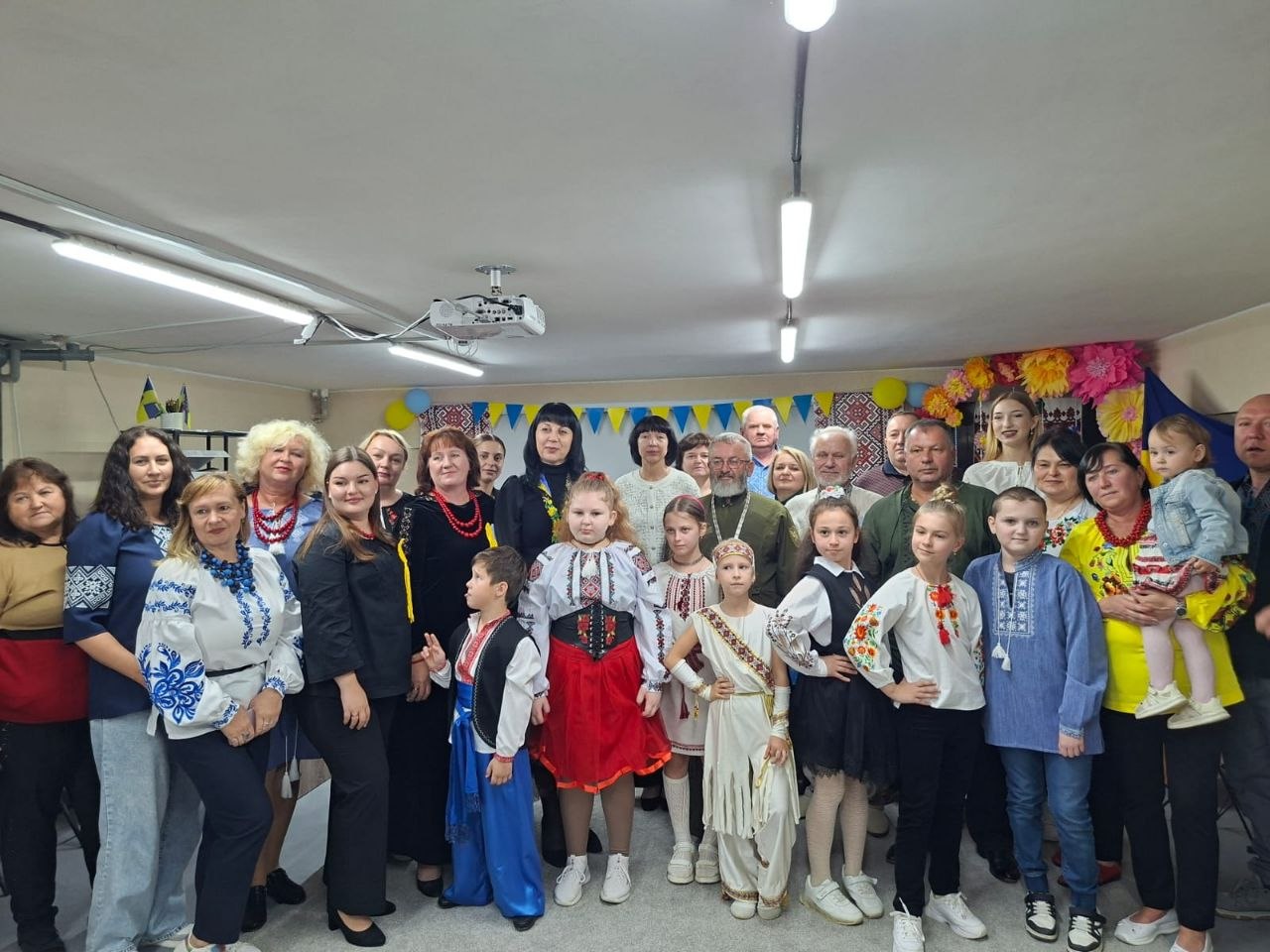 Спільна пам'ятна світлина всіх учасників, організаторів і гостей святкового заходу. Велика група дорослих і дітей у вишиваному вбранні вишикувалася у залі після концерту. У центрі стоять діти у сценічних костюмах, поруч представники духовенства та громади, а приміщення прикрашене гірляндами з прапорців, рушниками й українською символікою.