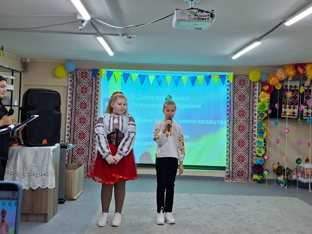 Дві дівчинки у вишиванках виступають перед глядачами. Дівчинка праворуч у темних штанях тримає мікрофон і декламує, поруч стоїть дівчинка у червоній спідниці та корсетці. На задньому плані транслюється тематична презентація до Дня захисників і захисниць України, обабіч якої розвішані ткані рушники з традиційним орнаментом і паперові квіти.