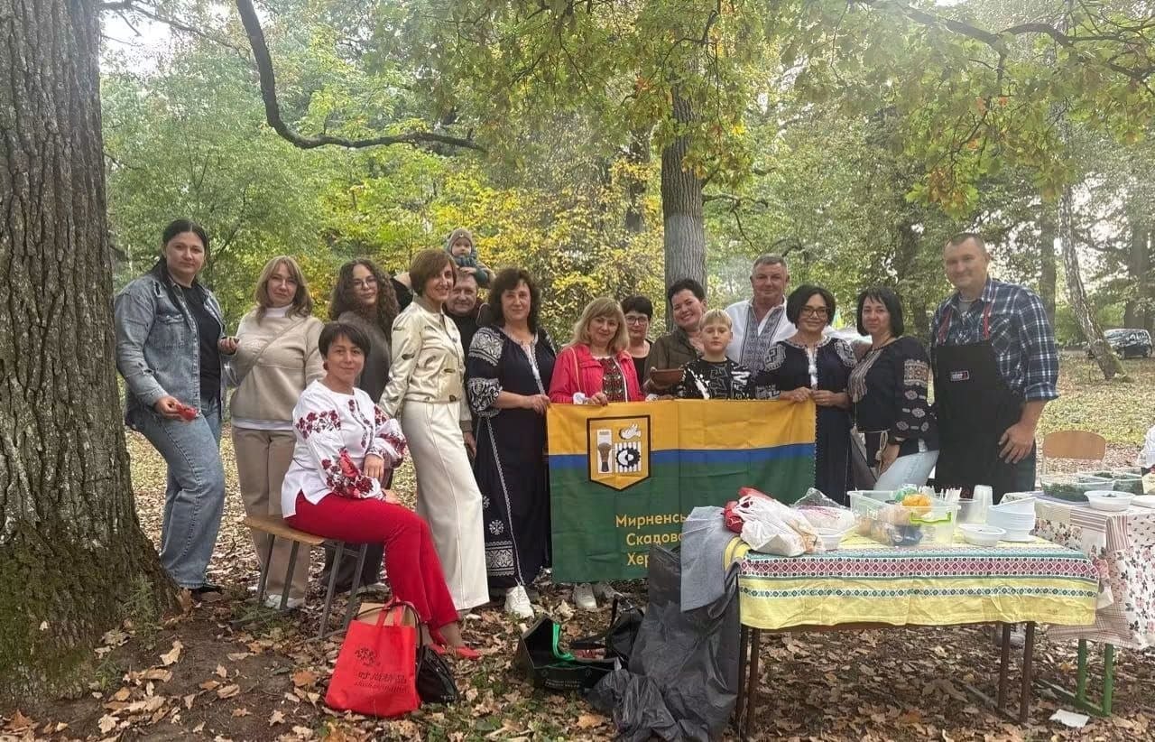 Група жителів і представників громади на виїзному заході серед осіннього дубового лісу. Учасники у вишиванках і повсякденному одязі зібралися навколо столу з польовим частуванням. По центру вони тримають горизонтальний прапор Мирненської селищної територіальної громади Скадовського району Херсонської області.
