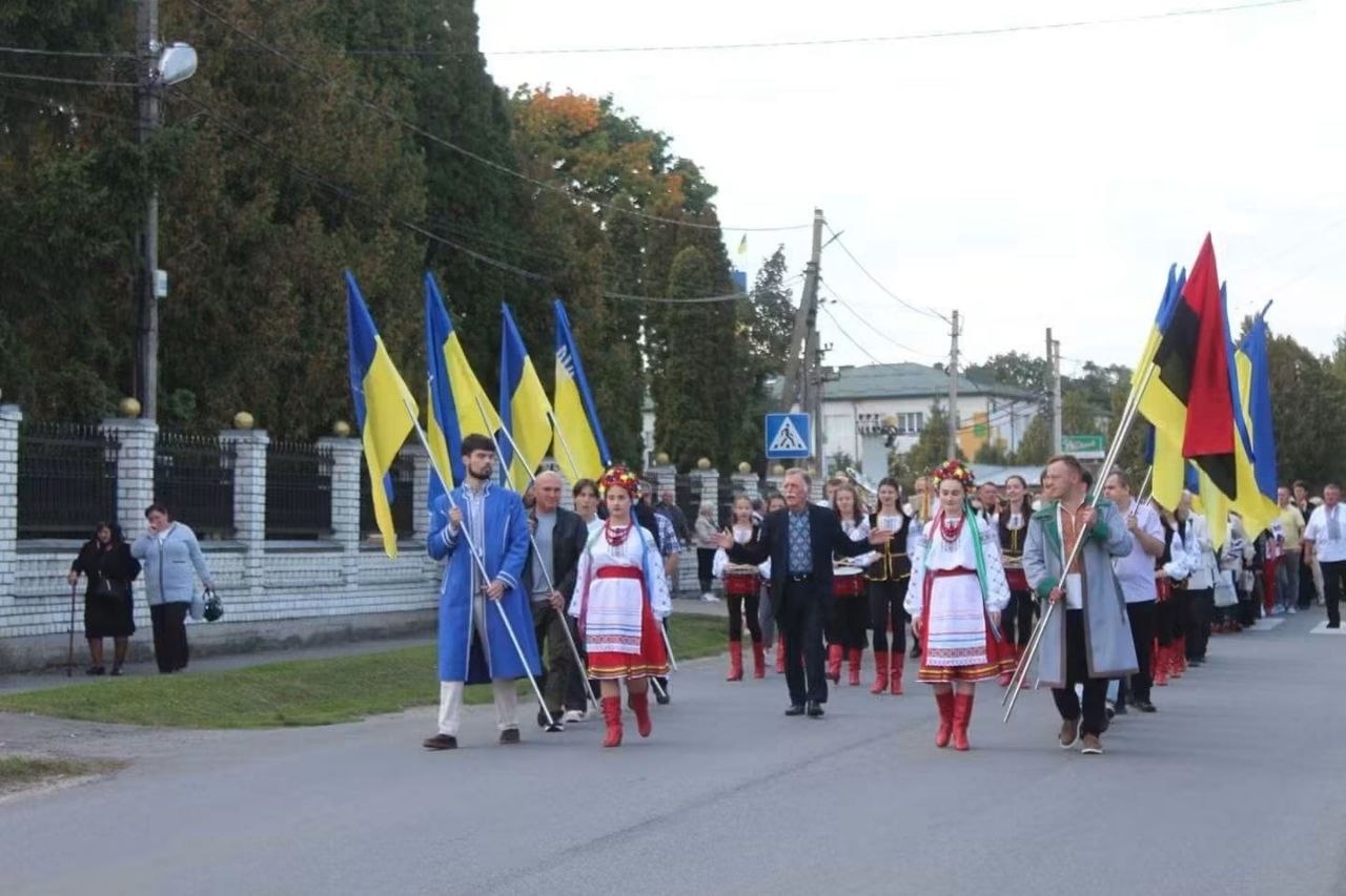 Святкова патріотична хода вулицею населеного пункту. Попереду колони йдуть юнаки та дівчата в традиційних українських строях і козацьких жупанах: ліворуч несуть великі синьо-жовті прапори України, праворуч — червоно-чорні та державні стяги. У центрі крокує чоловік у темному костюмі та вишиванці, поруч ідуть учасниці у вінках і вишитих сукнях. Позаду рухається організована колона людей на тлі зелених дерев і будинків.