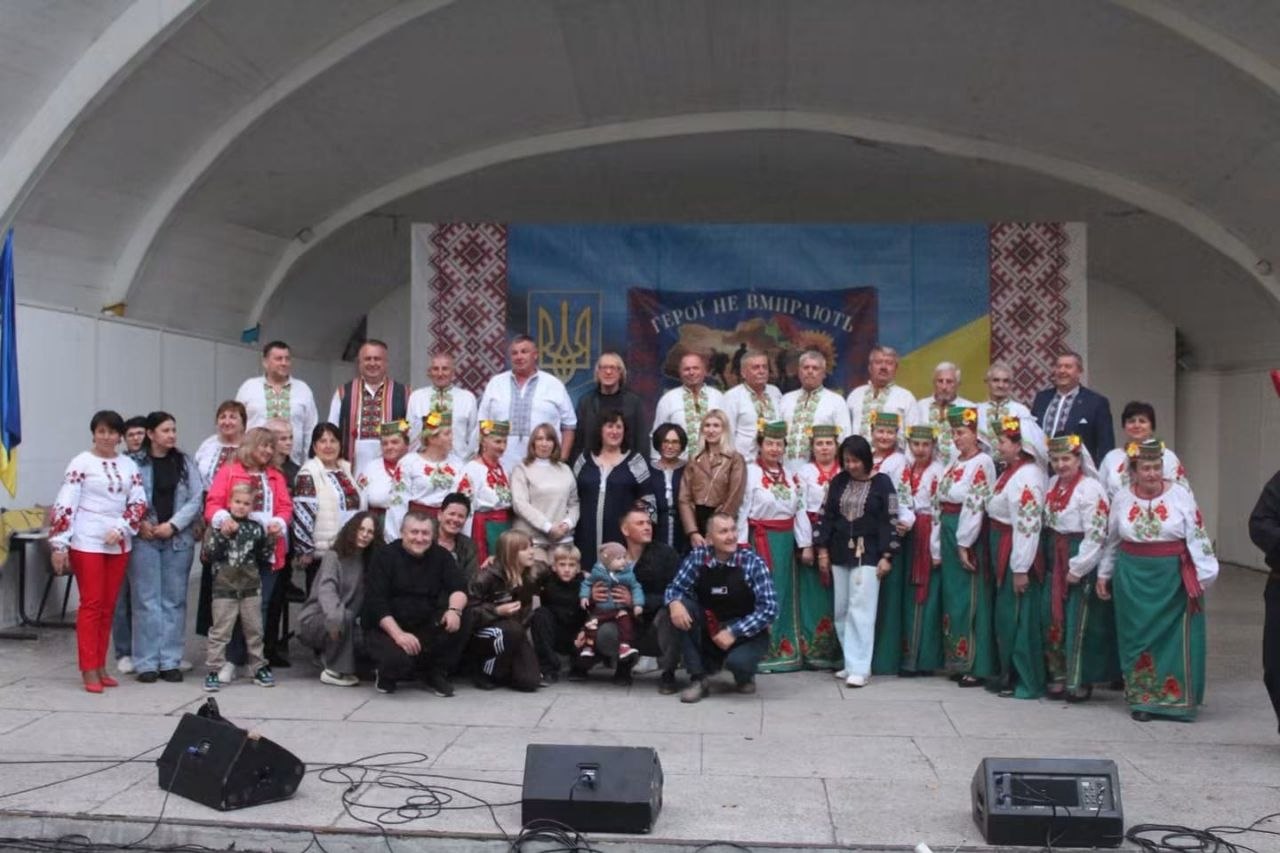 Велика спільна фотографія учасників фольклорного свята на напівкруглій критій сцені. Позаду стоять учасники хору та колективів у зелених спідницях із квітковими візерунками, вінках і вишиванках. Попереду розташувалися сім'ї з дітьми та організатори заходу. Задній план сцени прикрашений рушниками, патріотичним стендом «Герої не вмирають» та прапорами. На краю сцени встановлені сценічні монітори та звукове обладнання.