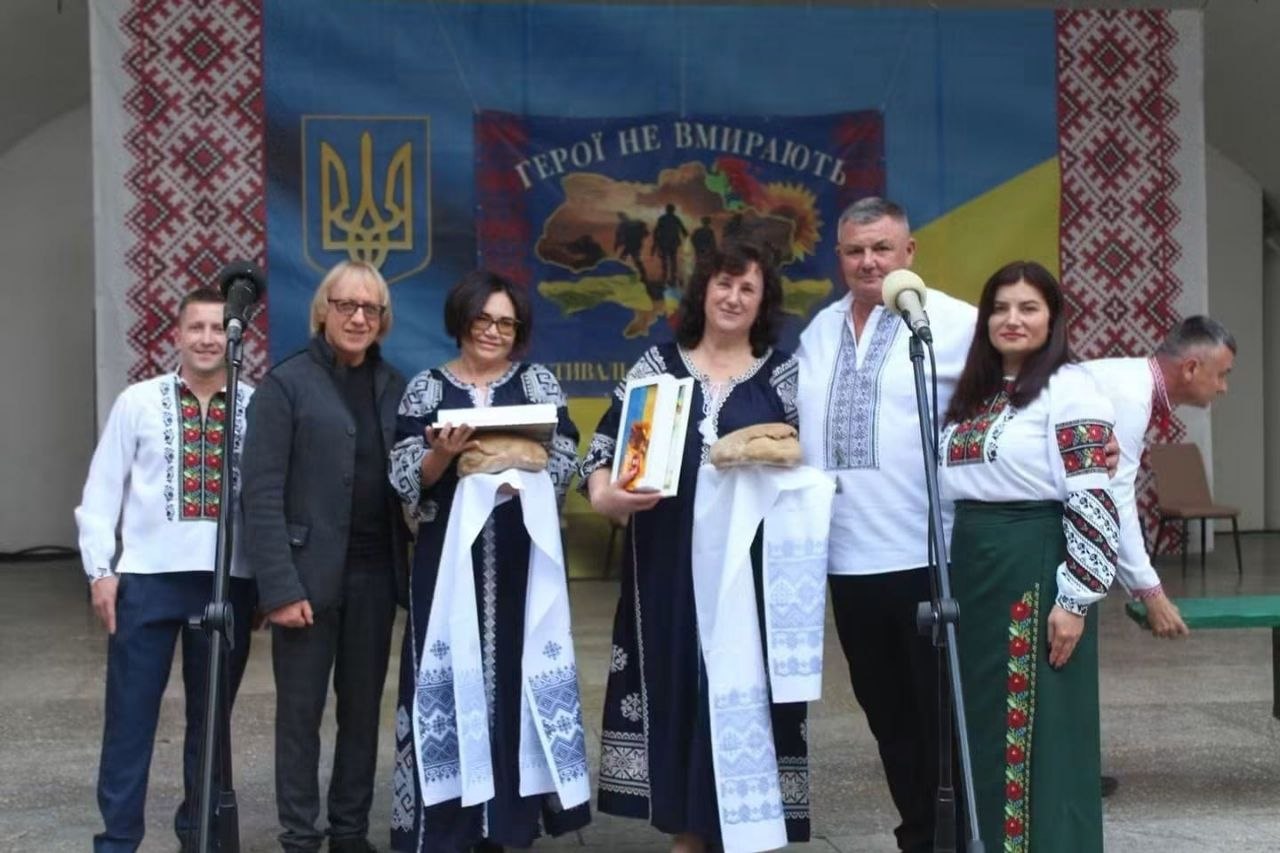 Урочистий момент вшанування на відкритій сцені. Шестеро учасників заходу в традиційних вишитих сорочках стоять біля мікрофонних стійок. Дві жінки по центру тримають на вишитих білих рушниках святкові короваї та пам'ятну книгу в синьо-жовтій обкладинці. Позаду встановлено банер із Тризубом, синьо-жовтим полотном та написом «ГЕРОЇ НЕ ВМИРАЮТЬ».