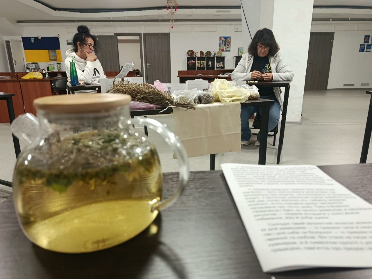 Затишна атмосфера майстер-класу з фітотерапії та створення ароматичних саше. На передньому плані на темному столі стоїть прозорий скляний чайник із завареним трав'яним чаєм золотистого відтінку та лежить розгорнута брошура з текстом. На задньому плані за партами сидять дві жінки: одна зосереджено перебирає засушені трави за столом з пучками полину та пакетиками рослин, інша читає методичні матеріали.
