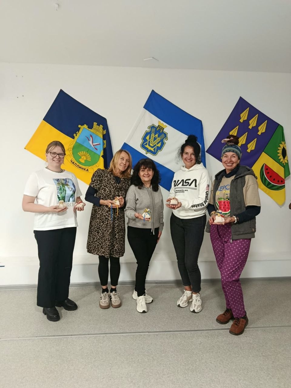 П'ять усміхнених учасниць заходу стоять у світлій кімнаті, тримаючи в руках власноруч створені трав'яні обереги та сувеніри. На стіні позаду них висять прапори: Державний Прапор України з гербом селища Чаплинка, синьо-білий прапор Херсонської області та темно-синій прапор із символікою соняшника, скіфських наконечників і скибки кавуна.