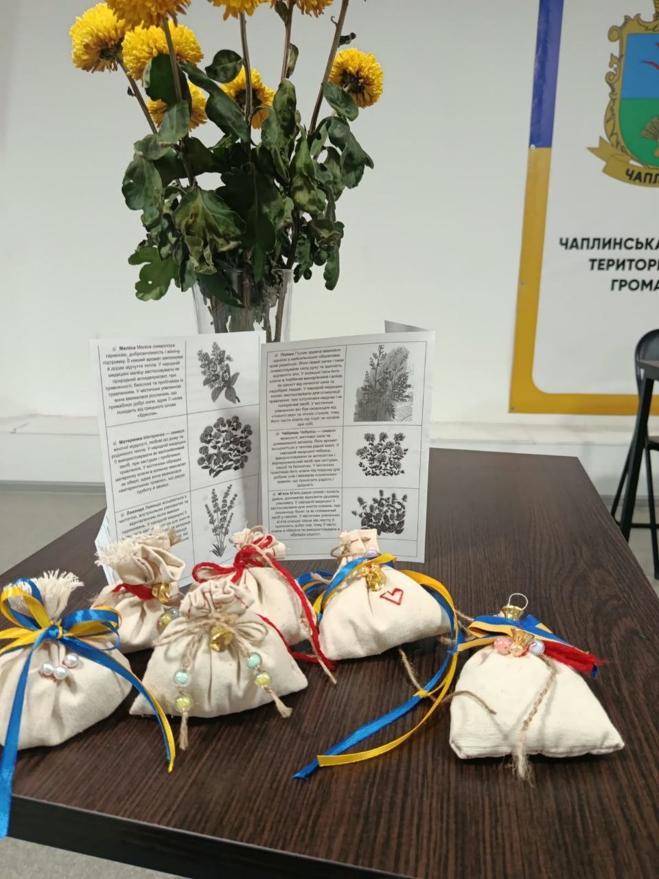 Чотири виготовлені власноруч лляні мішечки з цілющими травами, викладені в ряд на столі. Саше перев'язані джгутовими мотузками, синьо-жовтими та червоними стрічками й прикрашені декоративними намистинами та вишитим червоним сердечком. Позаду них стоїть розгорнута ілюстрована пам'ятка про властивості трав (меліса, материнка, лаванда, чебрець) та ваза з букетом жовтих хризантем біля банера Чаплинської громади