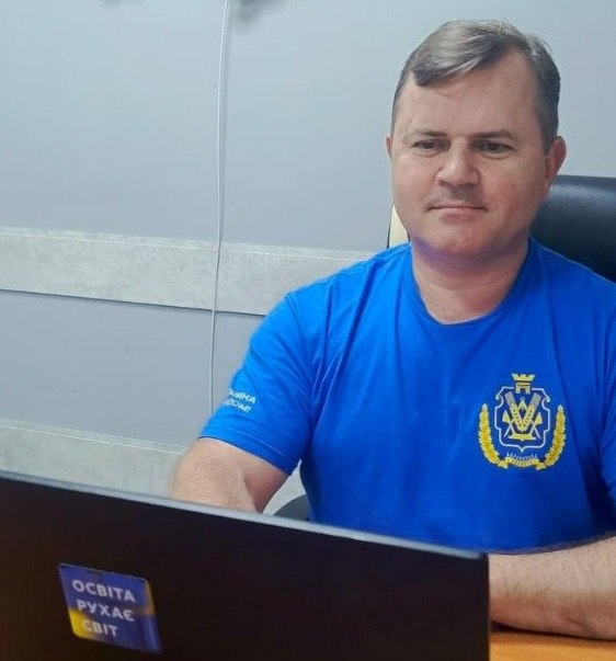 Заступник начальника Херсонської обласної військової адміністрації Володимир Клюцевський сидить за робочим столом у кабінеті під час онлайн-включення. Кадр зафіксований збоку (ракурс у три чверті).  Він одягнений у яскраво-синю футболку, на лівій стороні грудей якої нанесено великий золотаво-синій герб Херсонської області. Чоловік зосереджено дивиться на екран відкритого чорного ноутбука, що стоїть перед ним. На кришці ноутбука в лівому нижньому кутку наклеєно невеликий синьо-жовтий стікер із текстом «ОСВІТА РУХАЄ СВІТ». На задньому плані видно світло-сіру стіну кабінету та спинку офісного крісла.
