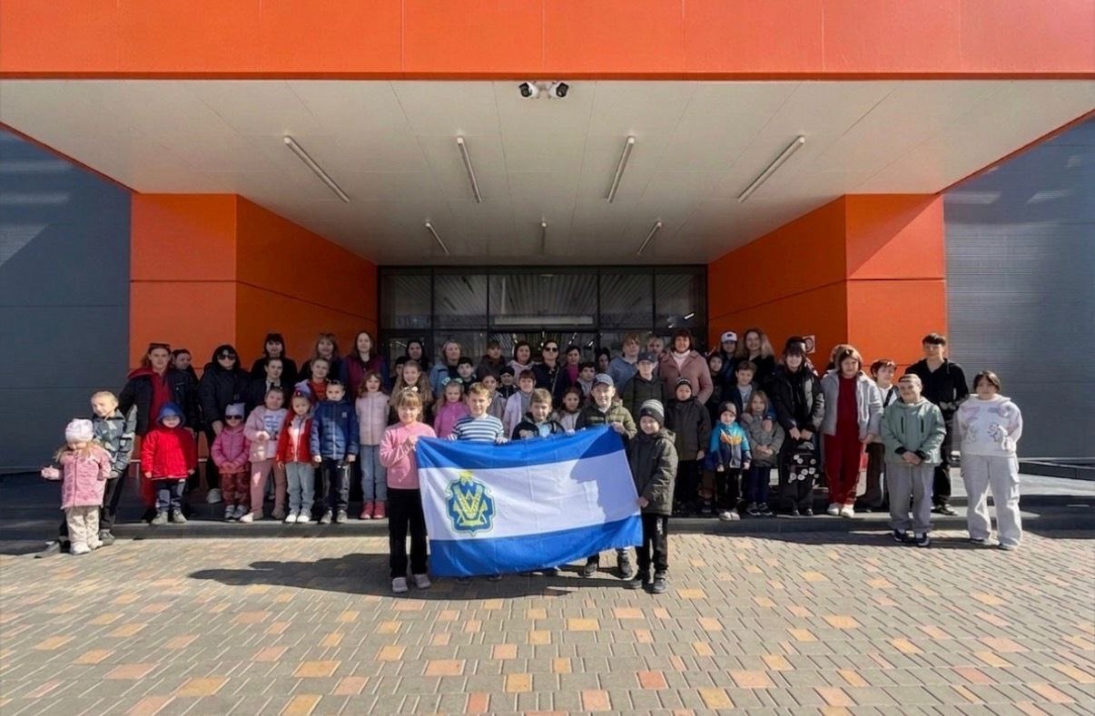 Велика група дітей різного віку та кілька дорослих стоять перед сучасним будинком з яскраво-помаранчевим фасадом. У центрі діти тримають великий синьо-білий прапор із золотистим гербом. День сонячний, усі дивляться в камеру.