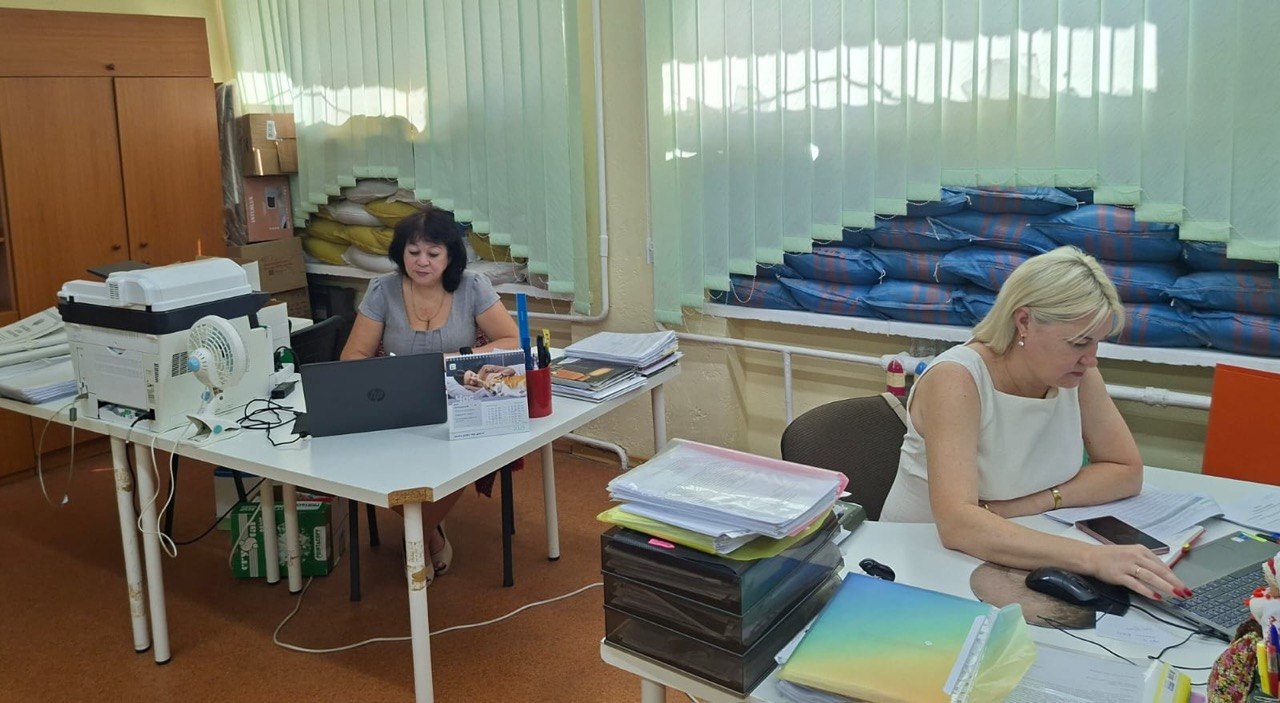 Робочий кабінет установи, де за двома окремими білими столами працюють дві жінки-співробітниці. Приміщення облаштоване з урахуванням безпекових вимог: нижні частини вікон завішені світло-зеленими вертикальними жалюзі, а підвіконня повністю закладені щільними рядами мішків із піском (жовтими ліворуч та синіми праворуч) для захисту від уламків. Ліворуч: жінка з темним волоссям у сірій блузі сидить за комп'ютерним столом. Перед нею відкритий чорний ноутбук HP, настільний календар із зображенням кота, канцелярія та стоси паперів. Поруч на краю столу встановлено великий білий багатофункціональний пристрій (принтер) та маленький білий вентилятор. У кутку за її спиною стоїть велика дерев'яна шафа. Праворуч: блондинка у білому топі без рукавів працює за своїм ноутбуком. Лівою рукою вона керує комп'ютерною мишею, а права лежить на клавіатурі. На столі перед нею розміщені папки з документами, кольоровий органайзер, лотки для паперів та мобільний телефон.