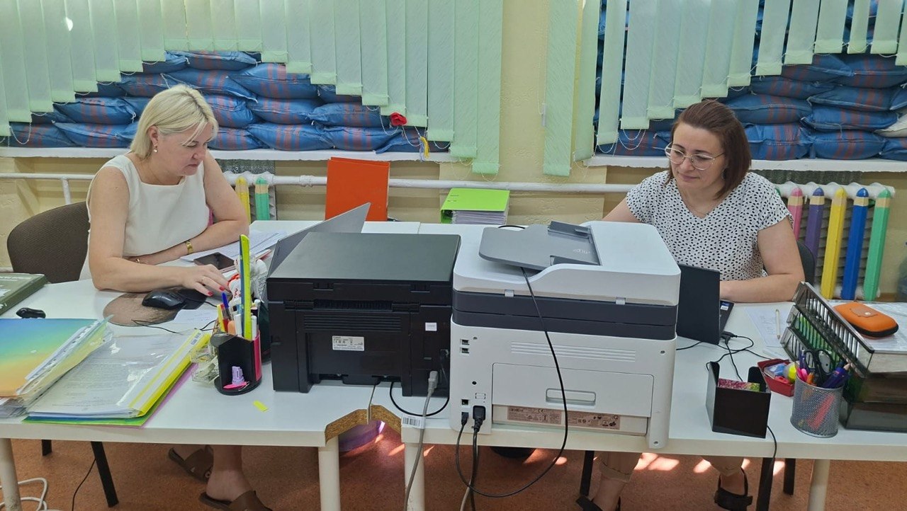 Робочий процес у кабінеті, зафіксований з іншого ракурсу (прямий план). Дві жінки-співробітниці сидять навпроти одна одної за здвоєним білим робочим столом, який розділяє оргтехніка. Уздовж усього вікна на задньому плані за світло-зеленими жалюзі видно захисні барикади з мішків із піском синього кольору. Ліворуч: блондинка у білому топі сидить на стільці, заклавши ногу на ногу. Вона зосереджено працює за ноутбуком, тримаючи руку на клавіатурі. Поруч із її робочою зоною лежать підготовлені теки з документами та підставка з ручками й маркерами. По центру: на стику столів задньою панеллю до камери встановлено два великі принтери — чорний ліворуч та біло-сірий багатофункціональний пристрій праворуч, до яких підключено кабелі живлення. Праворуч: жінка в окулярах та світлій блузці в дрібний чорний горошок сидить за своїм ноутбуком і посміхається. На її частині столу стоїть металева склянка з канцелярією, лотки та пенал для окулярів. Праворуч на стіні під вікном помітно декоративні елементи у вигляді великих різнокольорових олівців.