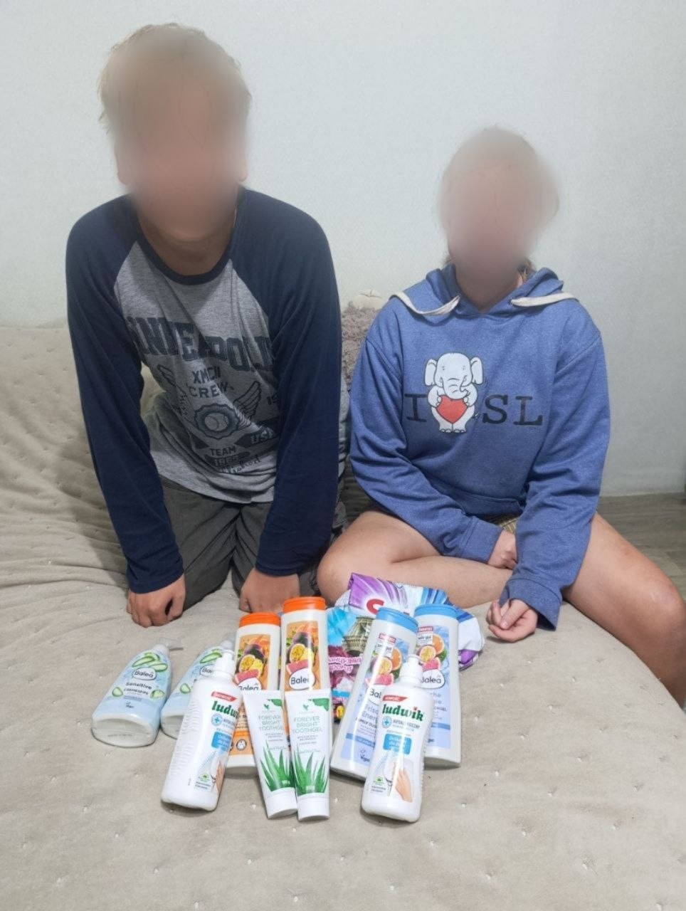 Двоє підлітків сидять на дивані зі світлим покривалом поруч із викладеними гігієнічними засобами. Перед ними лежать флакони рідкого крем-мила, шампуні Balea, засіб для прибирання Ludwik, тюбики зубної пасти з алое та упаковка порошку Savex. Обличчя обох дітей закриті розмиттям.