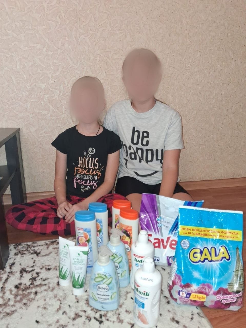 Дві дівчинки сидять на світлому килимі біля стіни, перед ними щільно розставлений набір побутової гігієни. Серед предметів гуманітарного набору: пачки прального порошку Gala та Savex, флакони мийних засобів Ludwik, рідке мило Balea Sensitive, зубні гелі та кілька пляшок шампунів. Обличчя дівчаток розмиті.
