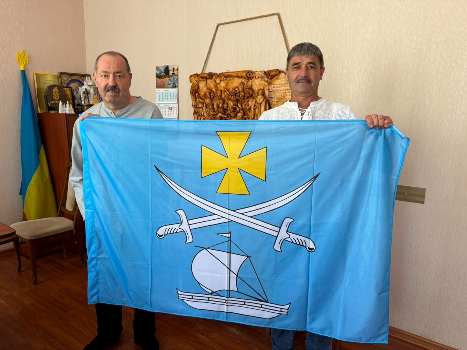 Два чоловіки середнього віку стоять у кабінеті та спільно тримають перед собою великий розгорнутий прапор Бериславського району (або міста Берислав) Херсонської області.  Полотно прапора має насичений блакитний (лазуровий) колір. У його центрі вертикально розміщено символіку:  Зверху — золотий розширений козацький хрест.  Посередині — дві схрещені срібні козацькі шаблі руків'ями донизу.  Знизу — білий козацький човен (чайка) під вітрилом, що пливе праворуч.  Чоловік ліворуч одягнений у світло-сіру кофту та чорні штани, має вуса. Чоловік праворуч одягнений у білу вишиванку з білим орнаментом та сині джинси, також має вуса. Обидва дивляться в об'єктив камери.  На задньому плані в лівому кутку приміщення стоїть державний прапор України на дерев'яному флагштоці із золотистим наконечником-тризубом, а поруч — дерев'яна тумба з нагородами, грамотами та кубками. На світлій бежевій стіні по центру зафіксовано настінний календар та велике декоративне дерев'яне панно з різьбленим художнім сюжетом. Підлога в кабінеті виконана з темно-коричневого лінолеуму або ламінату.