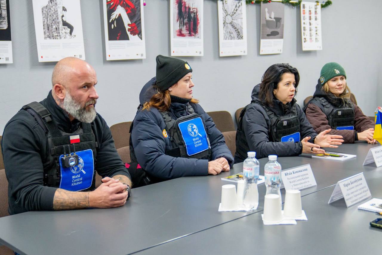 Група людей сидить за довгим столом під час офіційної зустрічі. Троє з них одягнені в чорні захисні жилети з синіми нашивками гуманітарної організації World Central Kitchen. На столі перед ними стоять пляшки з водою, паперові стаканчики та таблички з іменами учасників. На задньому плані на сірій стіні розвішані плакати з графічними ілюстраціями.