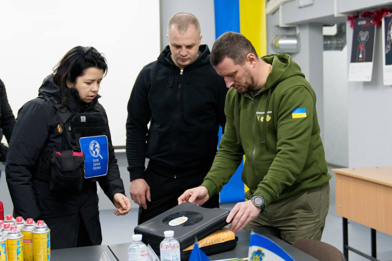 Три людини в приміщенні стоять біля столу та оглядають вміст відкритого чорного пластикового кейса. Чоловік праворуч одягнений у зелене худі з українським прапором на рукаві, а жінка ліворуч має на одязі шеврон гуманітарної організації World Central Kitchen. На столі поруч стоять пляшки з водою та балончики.