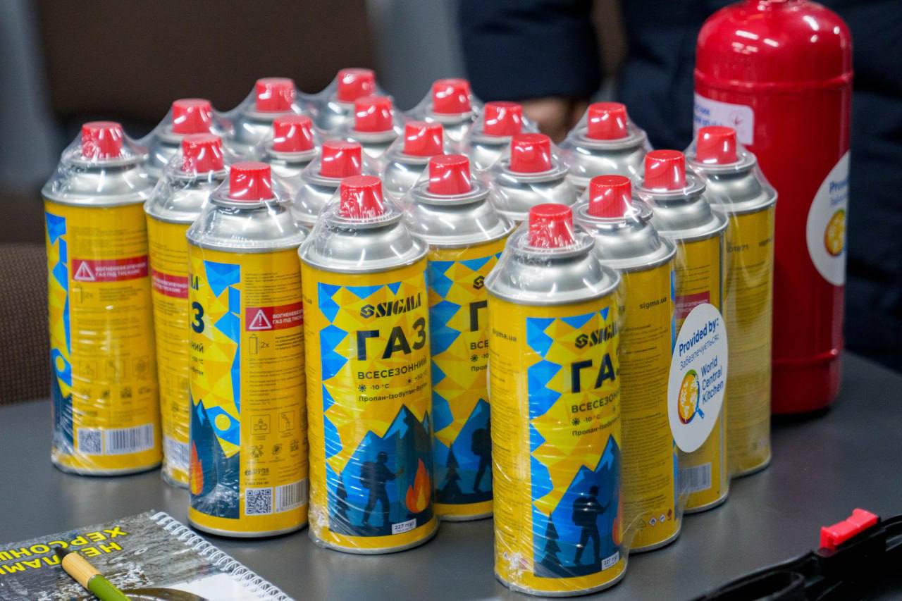 На столі стоїть велика упаковка нових газових балонів жовтого кольору в прозорій плівці. На етикетках балонів є напис «ГАЗ» та логотип бренду Sigma. Поруч на столі лежить блокнот із заголовком «Херсонщина» та стоїть червоний вогнегасник.