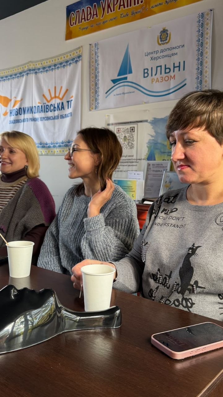 Учасниці заходу спілкуються за столом під час перерви. На передньому плані видно дівчину в сірій кофті з котячим принтом, поруч посміхається дівчина в окулярах і сірому в'язаному светрі. На столі стоять білі стаканчики з чаєм, лежить смартфон і знята пластикова маска. На стіні позаду розміщені банери Новомиколаївської ТГ Скадовського району та центру допомоги херсонцям «Вільні разом».