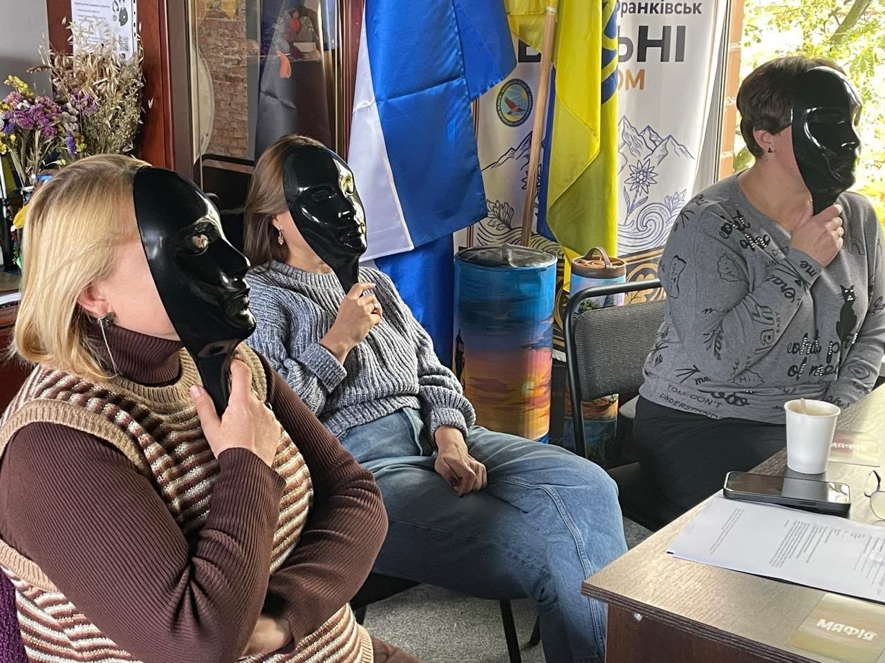 Троє учасниць сидять у ряд за столом під час гри в «Мафію», закривши обличчя чорними пластиковими масками на ручках. На столі перед ними лежать аркуші з правилами гри, напис «МАФІЯ» та стоїть паперовий стаканчик із напоєм. Позаду видно прапори, вертикальні банери гуманітарного хабу «Вільні разом м. Івано-Франківськ» і розписану тубус-гільзу біля стіни.