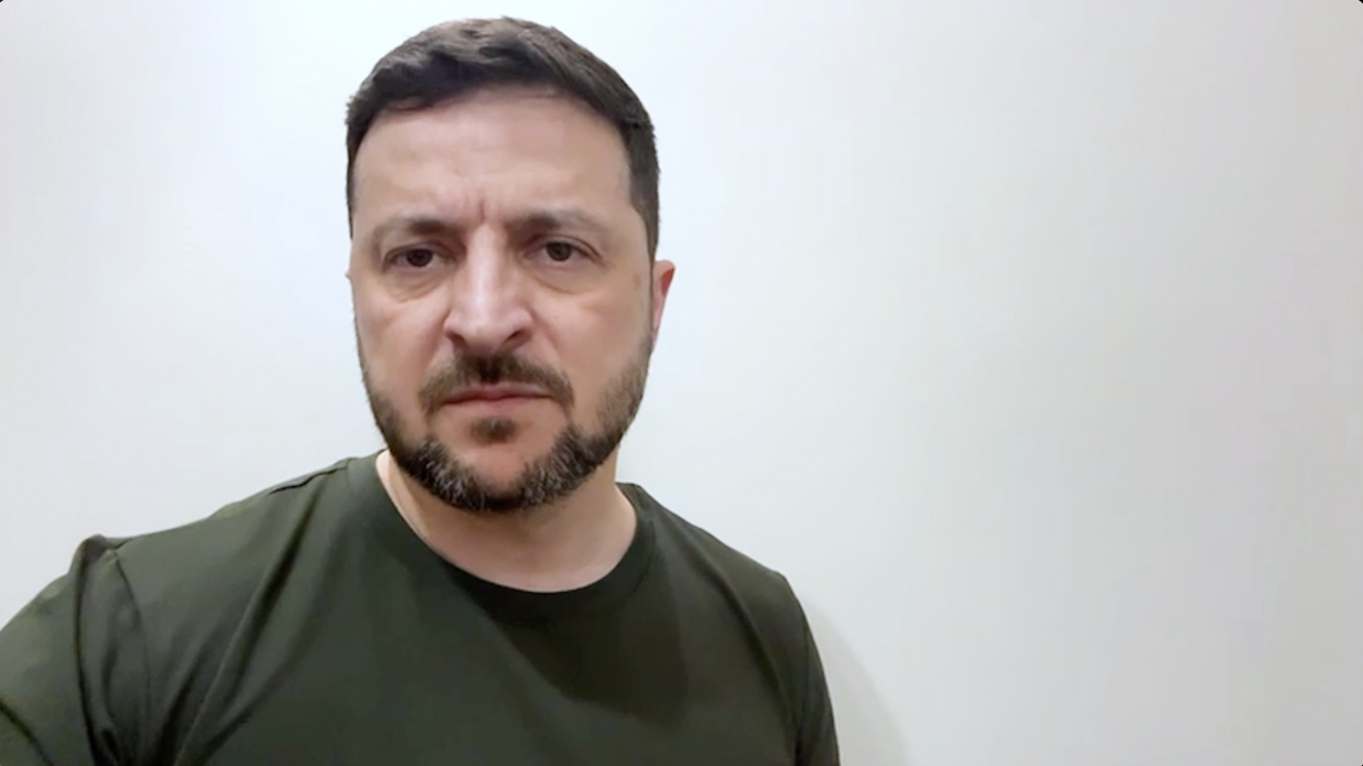 Крупний план Президента України Володимира Зеленського у футболці кольору хакі на тлі світлої стіни. Він дивиться прямо, з серйозним виразом обличчя.