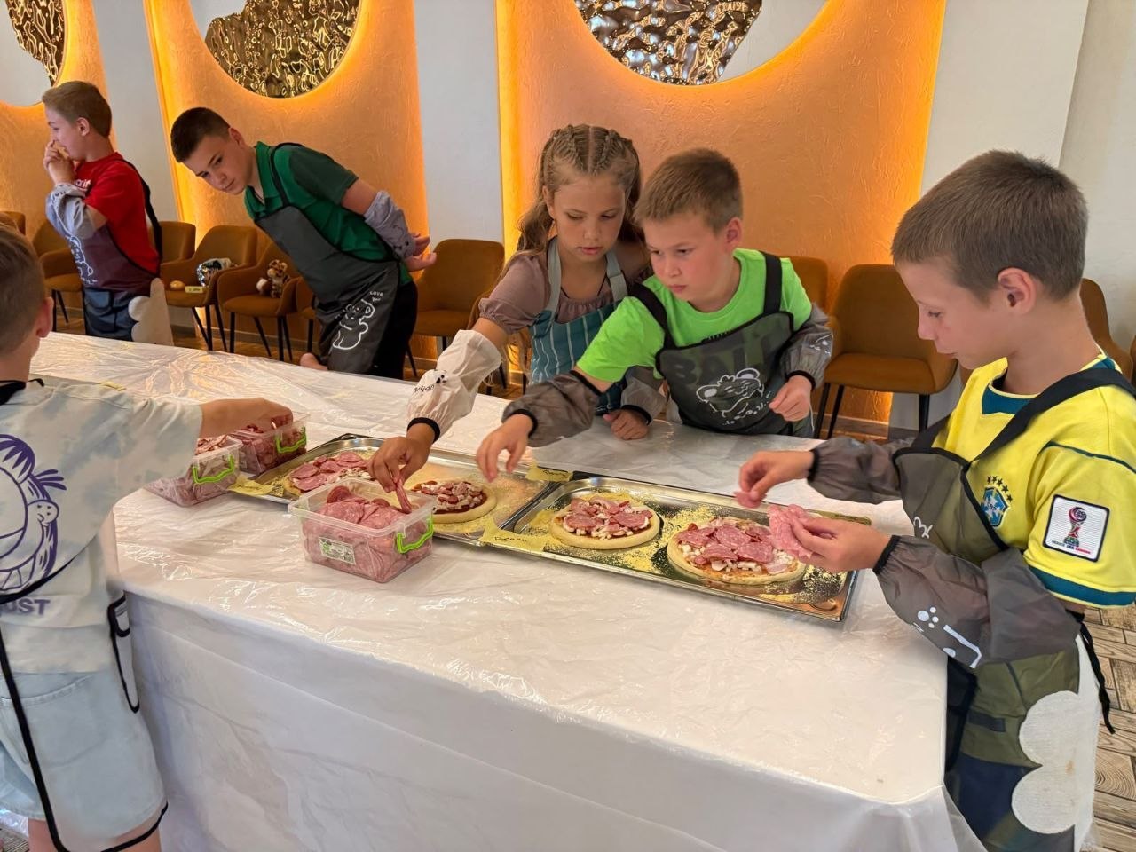 Четверо дітей за довгим столом, накритим білою захисною плівкою, захоплено готують піцу під час кулінарного заняття. Перед кожною дитиною на металевих деко лежать круглі заготовки з тіста, змащені соусом та посипані сиром. Діти у кулінарних фартухах та нарукавниках зосереджено викладають шматочки ковбаси та шинки із пластикових контейнерів, що стоять у центрі столу. На передньому плані праворуч хлопчик у жовто-синій футболці з логотипом збірної Бразилії з футболу акуратно кладе начинку на свою піцу. На задньому плані видно інших учасників майстер-класу та стіну з теплою помаранчевою підсвіткою і декоративними дзеркалами.