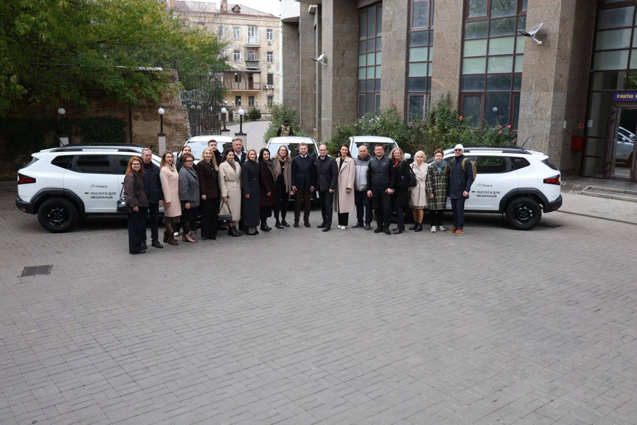 Групова фотографія близько двадцяти людей (чоловіків та жінок) у повсякденному одязі, які стоять на брукованій площі між двома білими позашляховиками (ймовірно, моделі Polaris). На бортах автомобілів видно логотип Polaris та напис