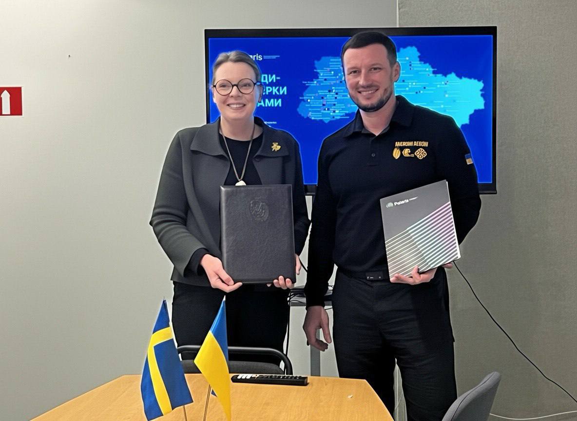 Двоє людей — жінка у чорному піджаку з окулярами та чоловік у темно-синій сорочці поло стоять, тримаючи документи (папки/буклети). Вони стоять перед великим екраном, на якому відображено карту України . На столі перед ними стоять мініатюрні прапори Швеції (синьо-жовтий хрест) та України (синьо-жовтий).