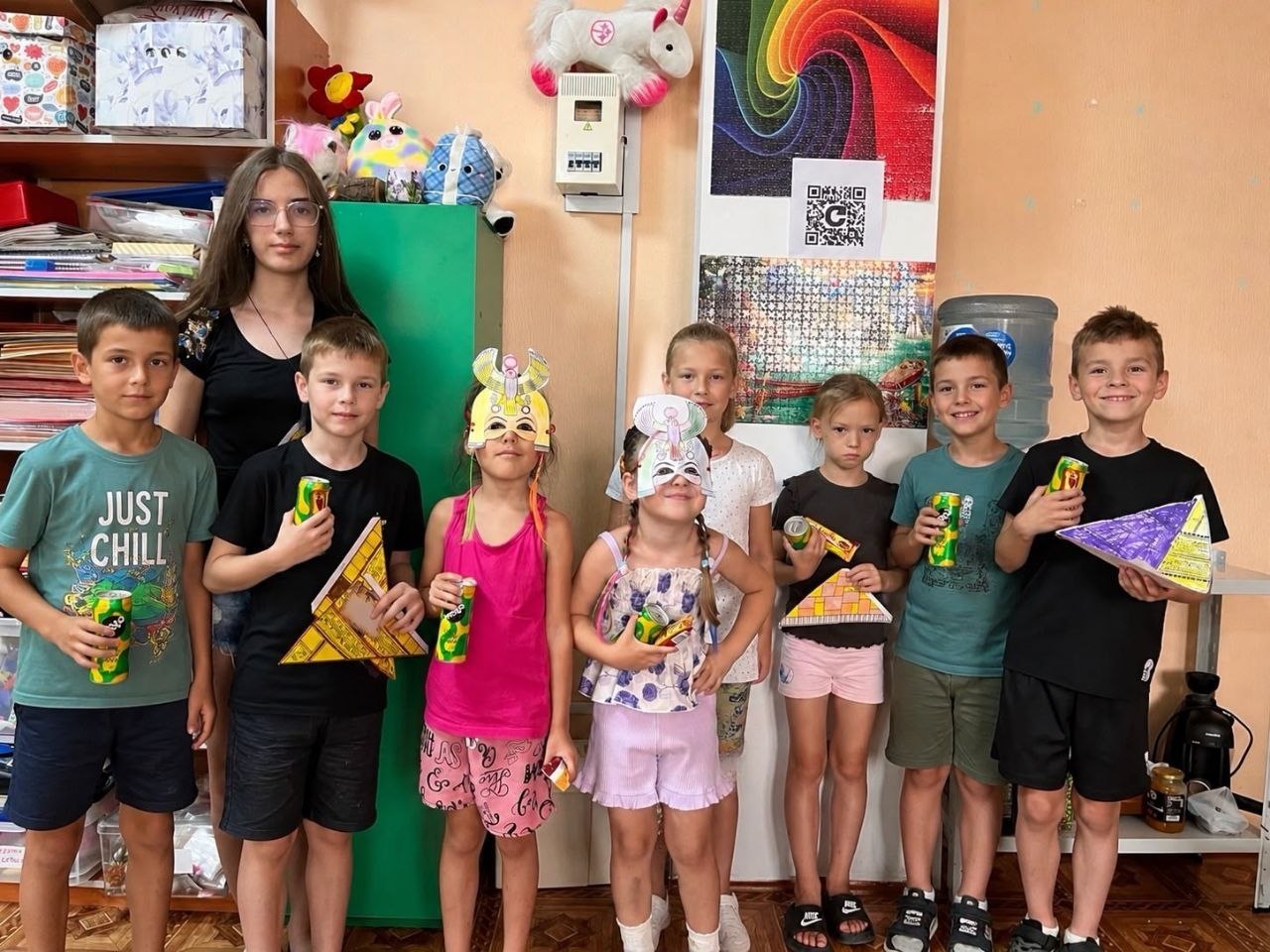 Групове фото восьми дітей та дівчини-підлітка, які вишикувалися в ряд у класі після завершення майстер-класу.  Діти тримають у руках свої готові вироби — зібрані з паперу об'ємні моделі єгипетських пірамід, кольорові маски фараонів та богів, а також банки з напоєм. Деякі дівчатка одягнули паперові єгипетські маски на голову. Задній план займає шафа з іграшками, стелажі з книгами та стіна з барвистою картиною-пазлом і QR-кодом.