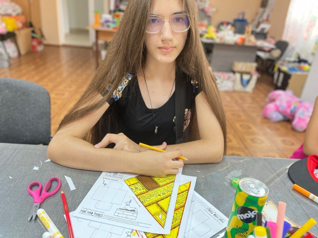 Дівчина-підліток у прозорих окулярах та чорній футболці із блискучим оздобленням на плечах сидить за великим сірим столом і дивується в об'єктив. Вона тримає в руці жовтий олівець.  Перед нею на столі розкладено роздруковані шаблони для об'ємних паперових саморобок із зображенням єгипетських пірамід та скульптур, рожеві ножиці, олівці, фломастери та зелена банка напою «Mojo». На задньому плані розмито видно інтер'єр дитячого закладу.