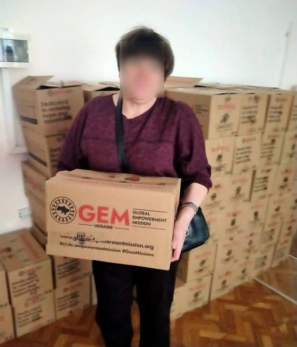 Жінка в темно-бордовій кофті тримає в руках велику картонну коробку гуманітарної допомоги від фонду GEM. На коробці нанесено логотип «GEM. GLOBAL EMPOWERMENT MISSION. UKRAINE» та вебaдресу організації. На задньому плані видно високу стопку аналогічних упакованих коробок, що заповнюють кімнату.