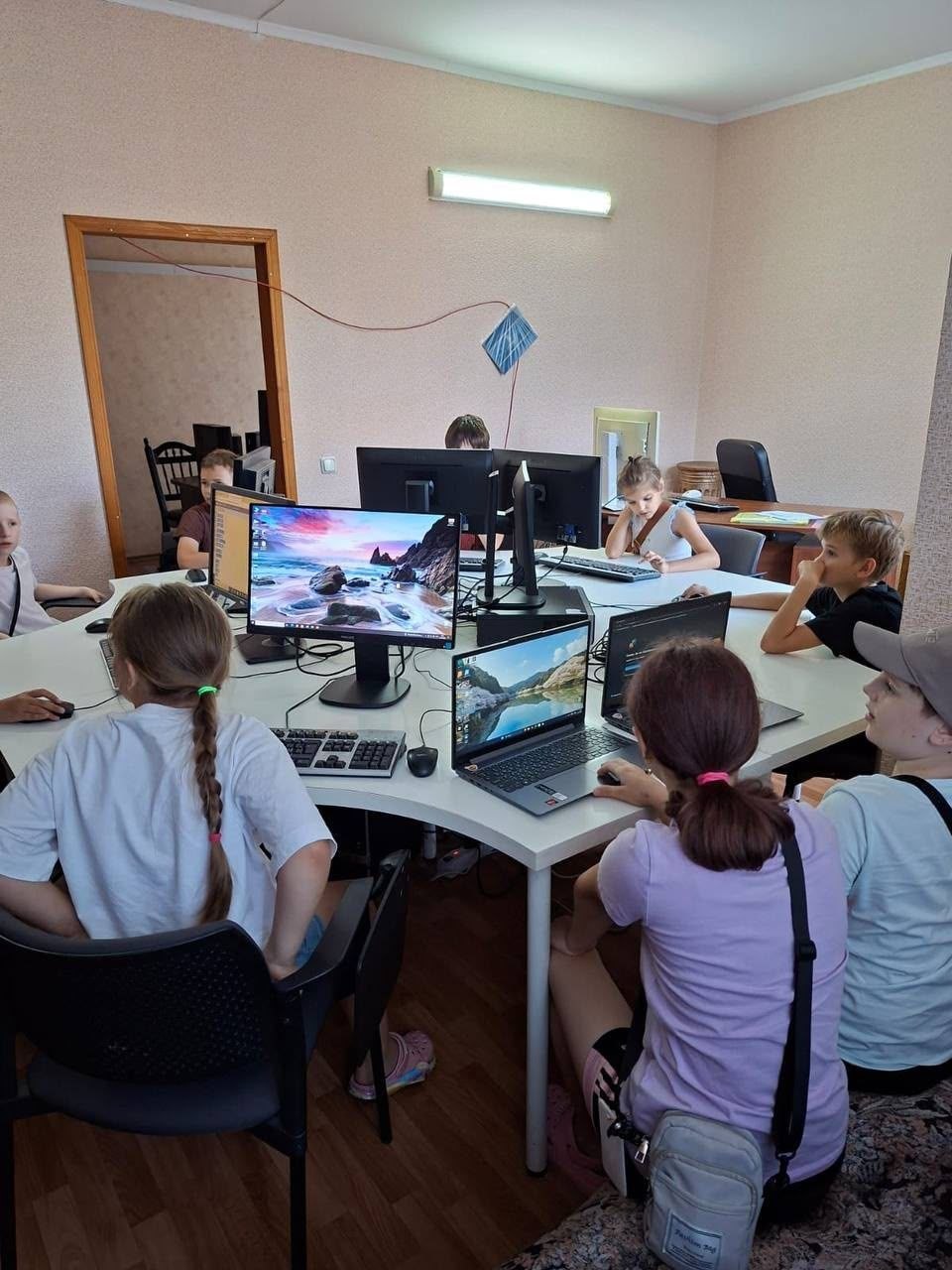 Діти та підлітки займаються в комп'ютерному класі за великим білим закругленим столом. Сім учасників сидять за моніторами та ноутбуками, вивчаючи матеріали чи виконуючи завдання. На екранах моніторів видно робочі столи та пейзажні шпалери. Приміщення освітлене люмінесцентною лампою, стіни персикового кольору, а на підлозі викладено лінолеум під паркет.