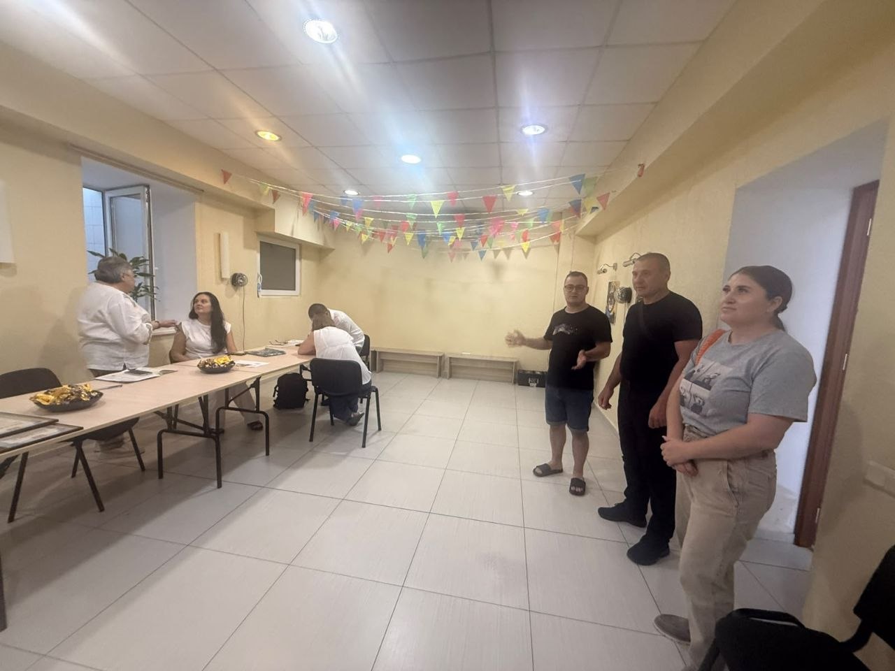 Загальний план робочої зустрічі у світлому просторому приміщенні з бежевими стінами та кахляною підлогою. Під стелею розтягнуто святкові гірлянди з кольоровими прапорцями.  Ліворуч за довгим столом сидять троє людей, одна жінка стоїть поруч та спілкується з присутніми. На столі розкладено документи та вази із солодощами. Праворуч стоїть група з трьох осіб: чоловік у чорній футболці та шортах, який жестикулює під час розмови, чоловік у чорному одязі та жінка в сірій футболці й бежевих штанах, яка уважно слухає.