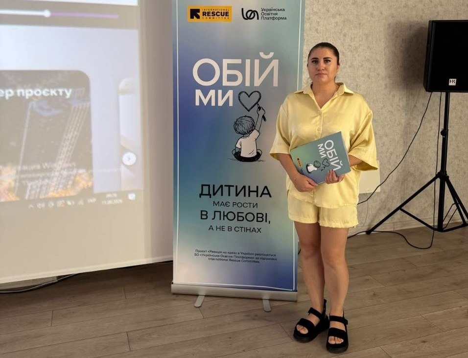 Дівчина у світло-жовтому костюмі з сорочки та шортів стоїть біля брендованого баннера проєкту «Обійми» і тримає в руках робочий зошит. На синьо-білому баннері поруч із нею розміщено логотипи International Rescue Committee та БО «Українська Освітня Платформа», назву проєкту «Обійми» з малюнком дитини, а також слоган: «Дитина має рости в любові, а не в стінах». Ліворуч у кадр потрапляє частина екрана з проекцією презентації, а праворуч — аудіоколонка на штативі.
