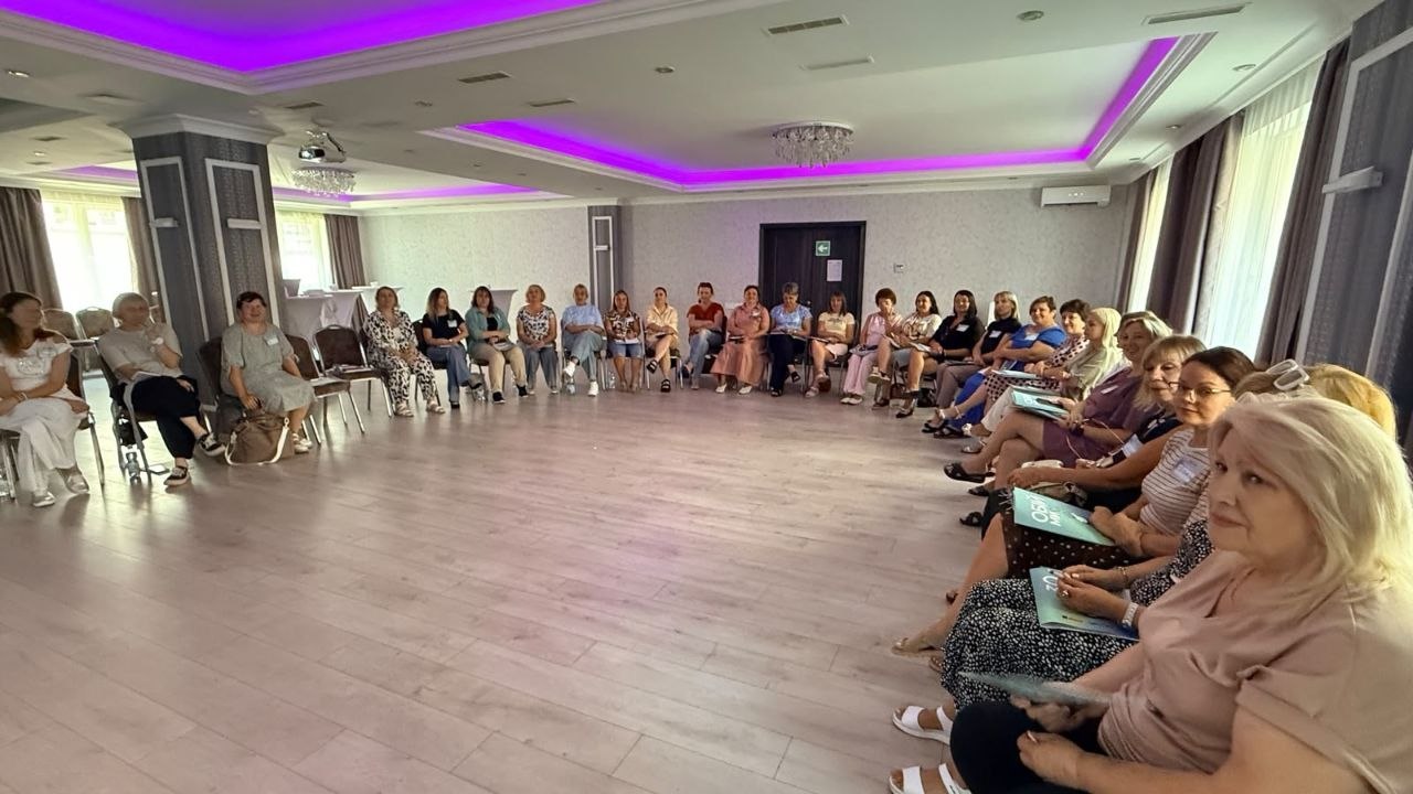 Загальний план інтерактивного заходу або тренінгу в просторій залі з фіолетовим неоновим підсвічуванням на стелі. Близько трьох десятків жінок сидять на стільцях, розставлених великим півколом уздовж стін. На передньому плані праворуч виділяються учасниці з робочими зошитами та матеріалами проєкту «Обійми» в руках. Жінки уважно слухають модератора та залучені до спільного обговорення.