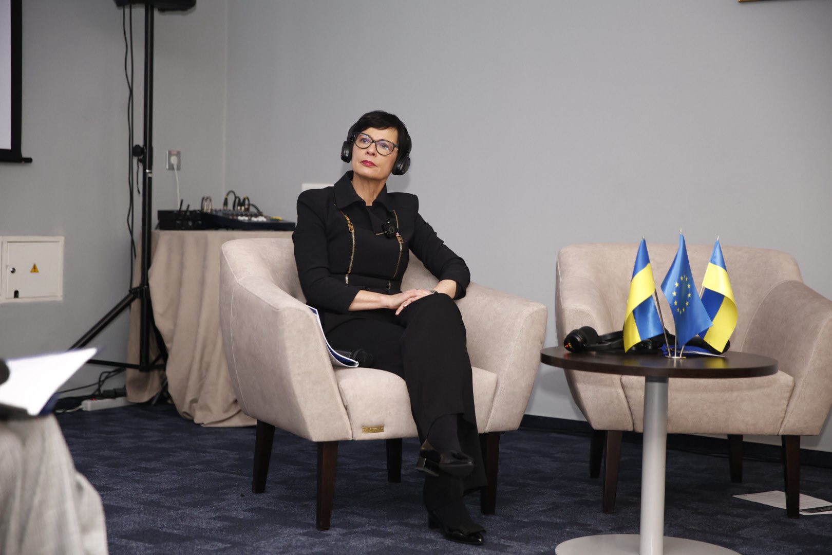 Жінка в окулярах та чорному офіційному костюмі сидить у світлому кріслі під час конференції. Вона в навушниках для синхронного перекладу та дивиться вгору. Поруч на столі розміщені прапорці України та Європейського Союзу.