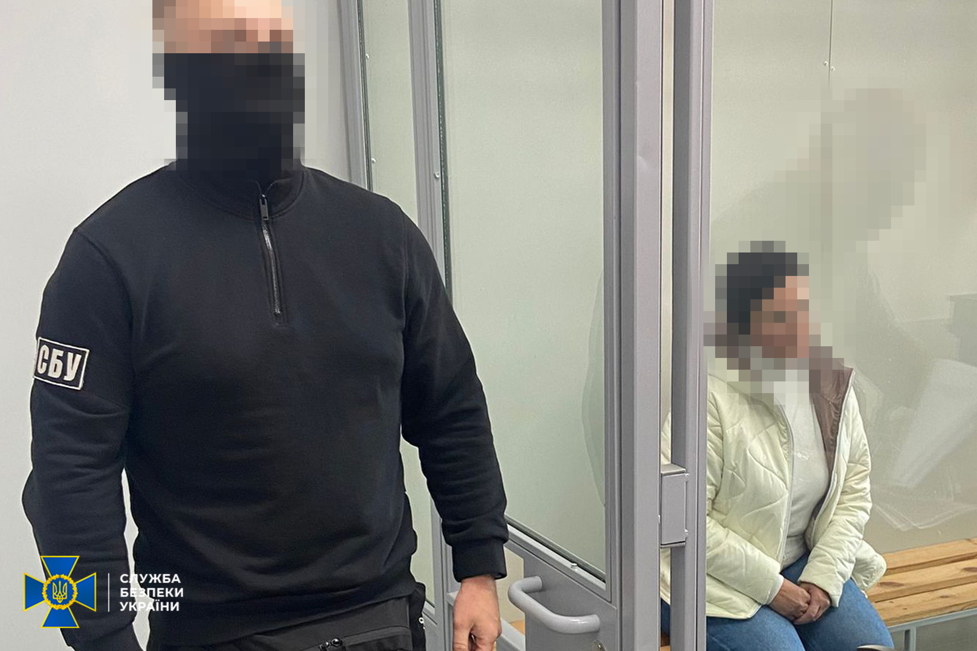 Фотографія із зали суду. На передньому плані ліворуч стоїть співробітник СБУ у чорній кофті на блискавці та з шевроном «СБУ» на рукаві. Його обличчя приховане за допомогою піксельного розмиття. Праворуч, за прозорим захисним склом кабіни для підсудних, на дерев'яній лаві сидить жінка у світлій куртці та синіх джинсах. Її обличчя також розмите. У лівому нижньому кутку кадру розміщено офіційний логотип Служби безпеки України.