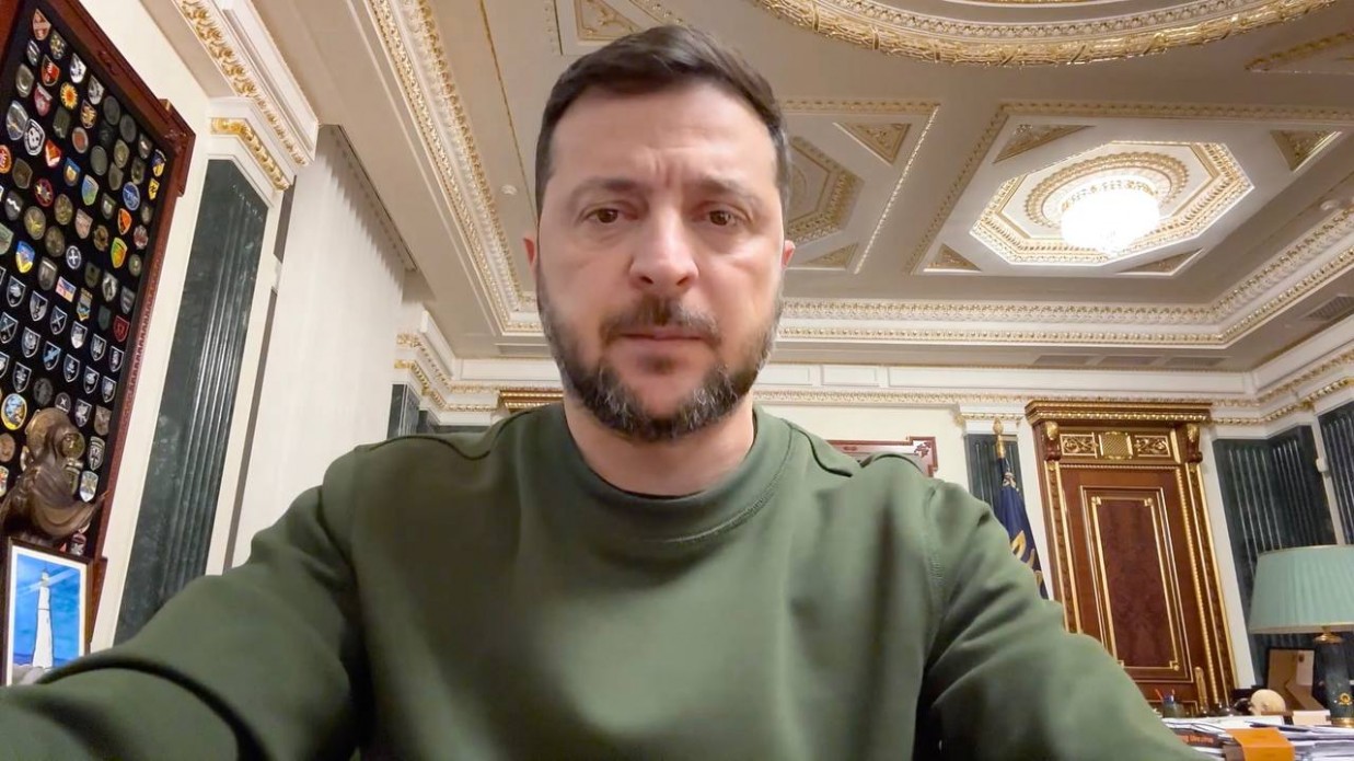 Портрет президента України Володимира Зеленського, який звертається до аудиторії. Він одягнений у свій звичний темно-зелений (оливковий) світшот і дивиться прямо в камеру. На задньому плані, ймовірно, його робочий кабінет: ліворуч на стіні велика вертикальна панель, повністю завішана медалями, значками та шевронами; праворуч — різьблені дерев'яні двері та класичний письмовий стіл з лампами, а стеля прикрашена декоративними ліпними елементами з позолотою.