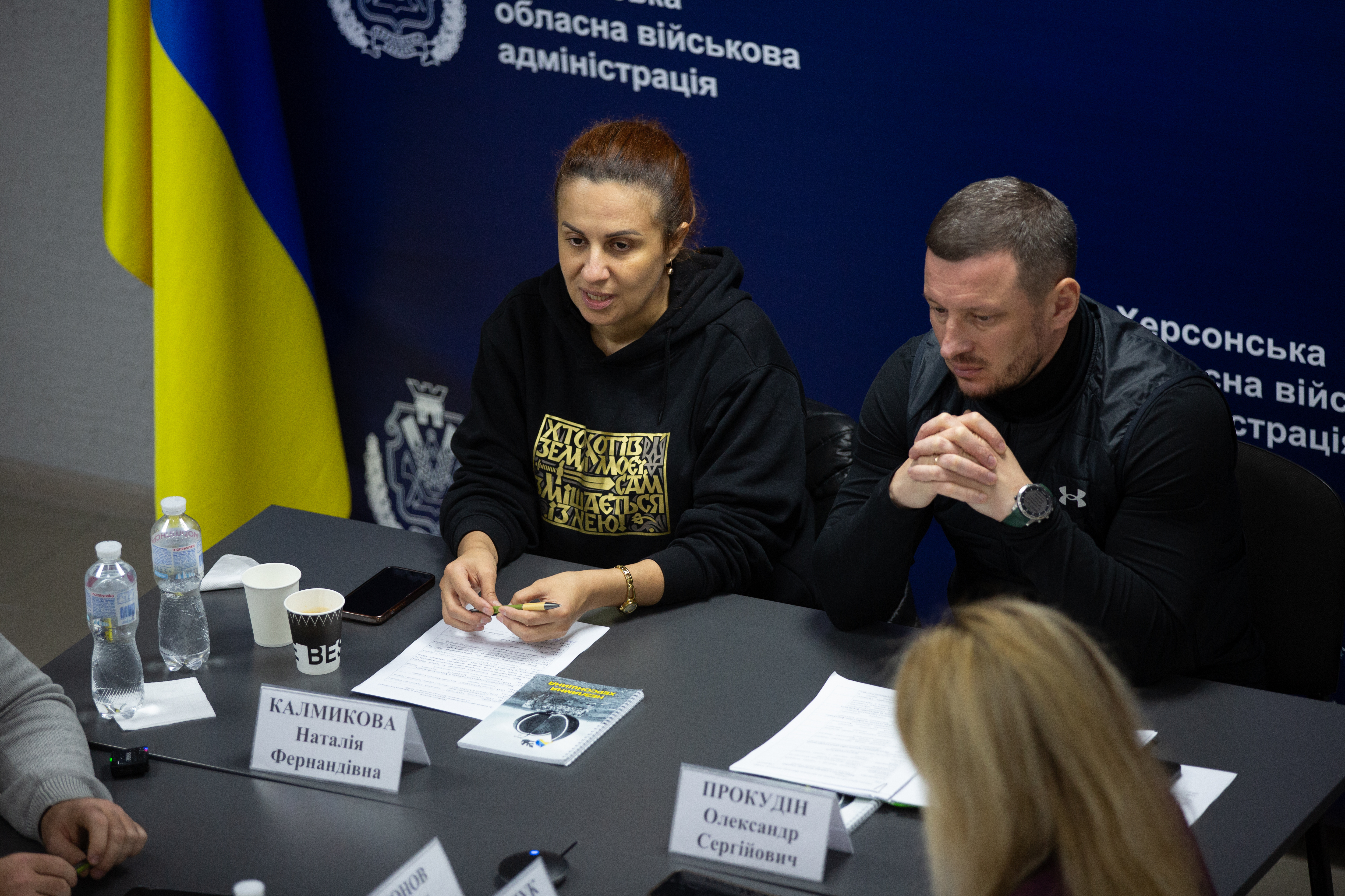Дві людини, ймовірно, чиновники, сидять за столом під час зустрічі. Жінка ліворуч у чорній кофті розмовляє, а чоловік праворуч у чорній спортивній кофті уважно слухає. На столі видно таблички з іменами. На задньому плані - прапор України та напис: Херсонська військова адміністрація.
