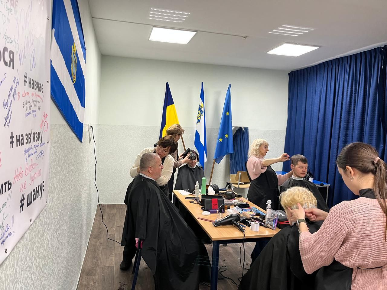 У світлому приміщенні майстри роблять стрижки чоловікам та жінкам. На столі розкладено професійні інструменти, а на фоні розташовані державні та регіональні прапори поруч із прапором ЄС.