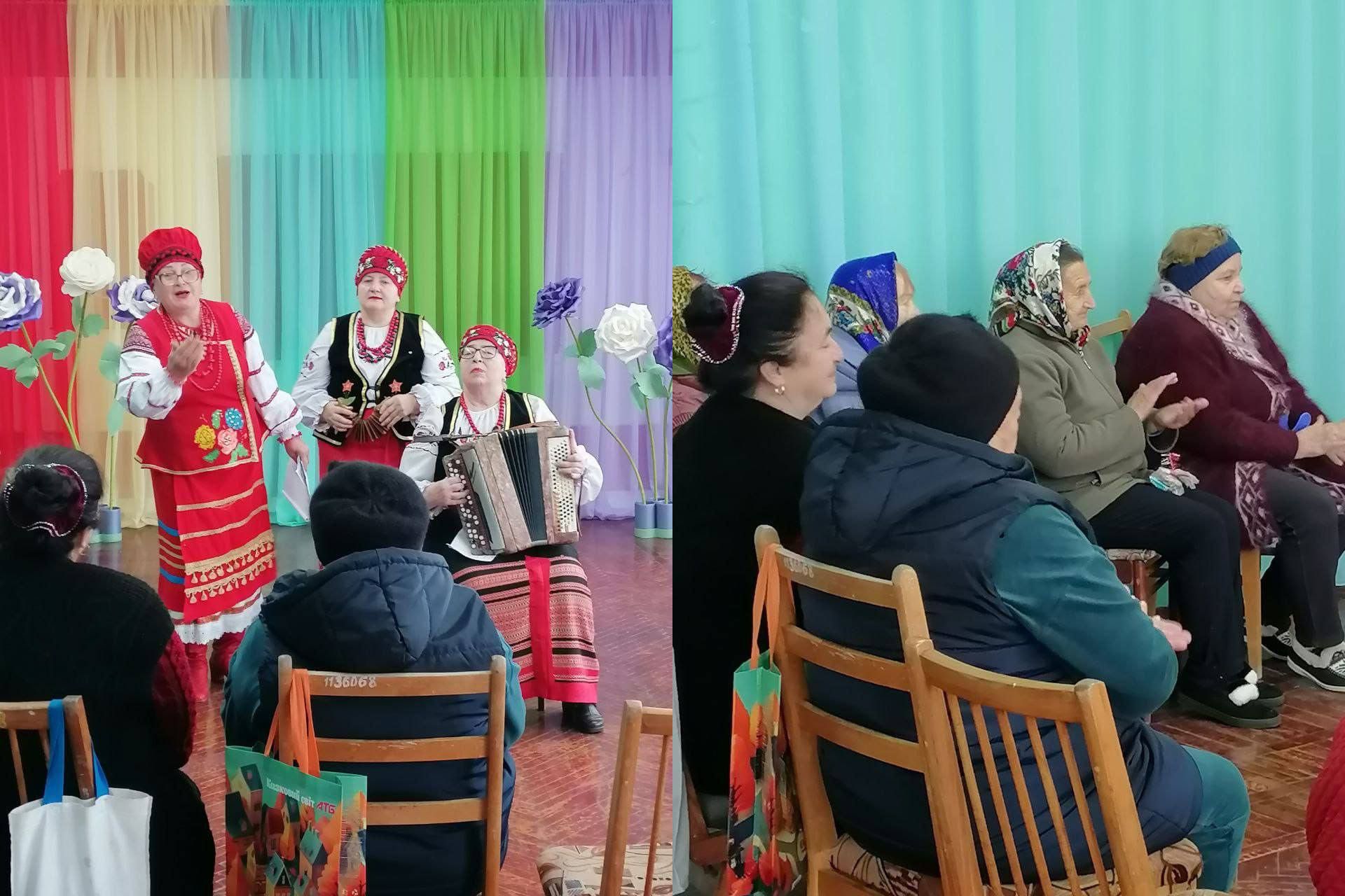 Сцена з культурного заходу: праворуч видно глядачів, переважно літніх жінок, які сидять і слухають. Зліва на сцені, прикрашеній вертикальними різнокольоровими шторами (червоний, бірюзовий, зелений, фіолетовий) та великими декоративними квітами, виступають три жінки в українських народних костюмах. Жінка в центрі грає на гармошці (баяні), а дві інші співають. На передньому плані видно спини глядачів.