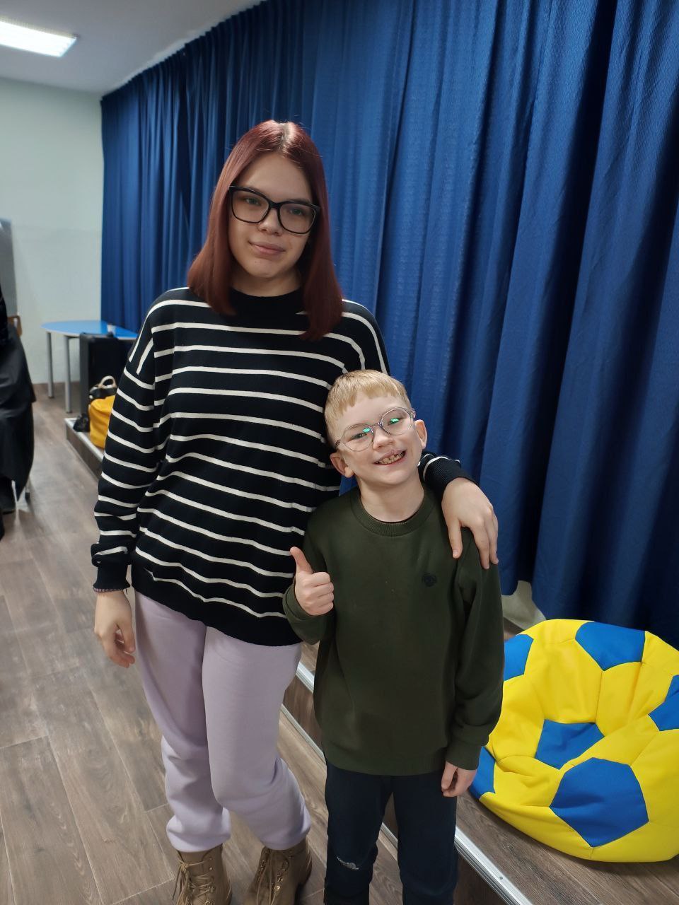 Дівчина в смугастому светрі обіймає за плечі хлопчика в окулярах, який показує великий палець вгору. Вони стоять у приміщенні на фоні синіх штор, поруч лежить крісло-мішок синьо-жовтого кольору.