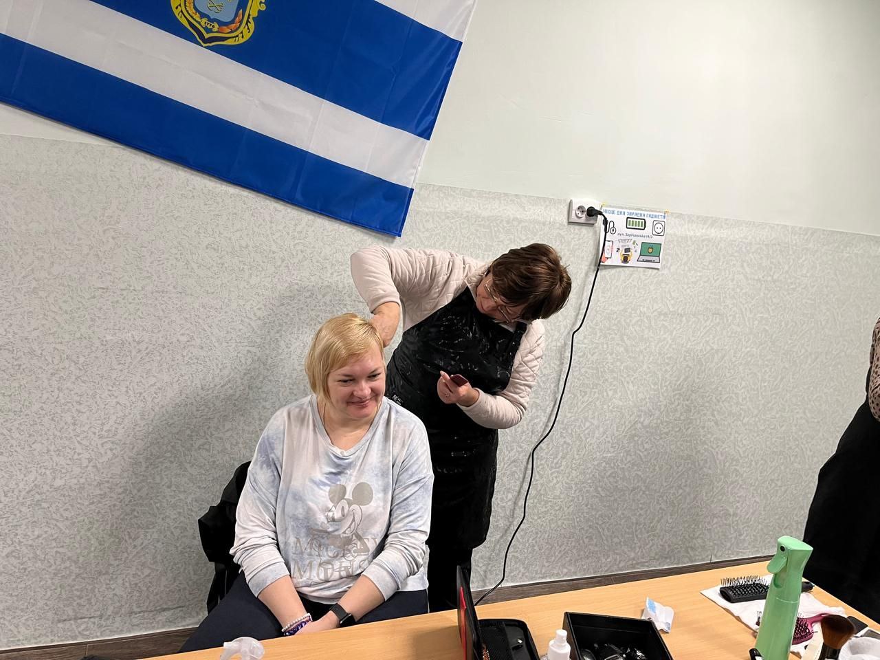 Перукар працює над зачіскою жінки, яка сидить перед столом з інструментами. Над ними на стіні закріплено синьо-білий прапор із гербом та інформаційну табличку про місце для зарядки гаджетів.