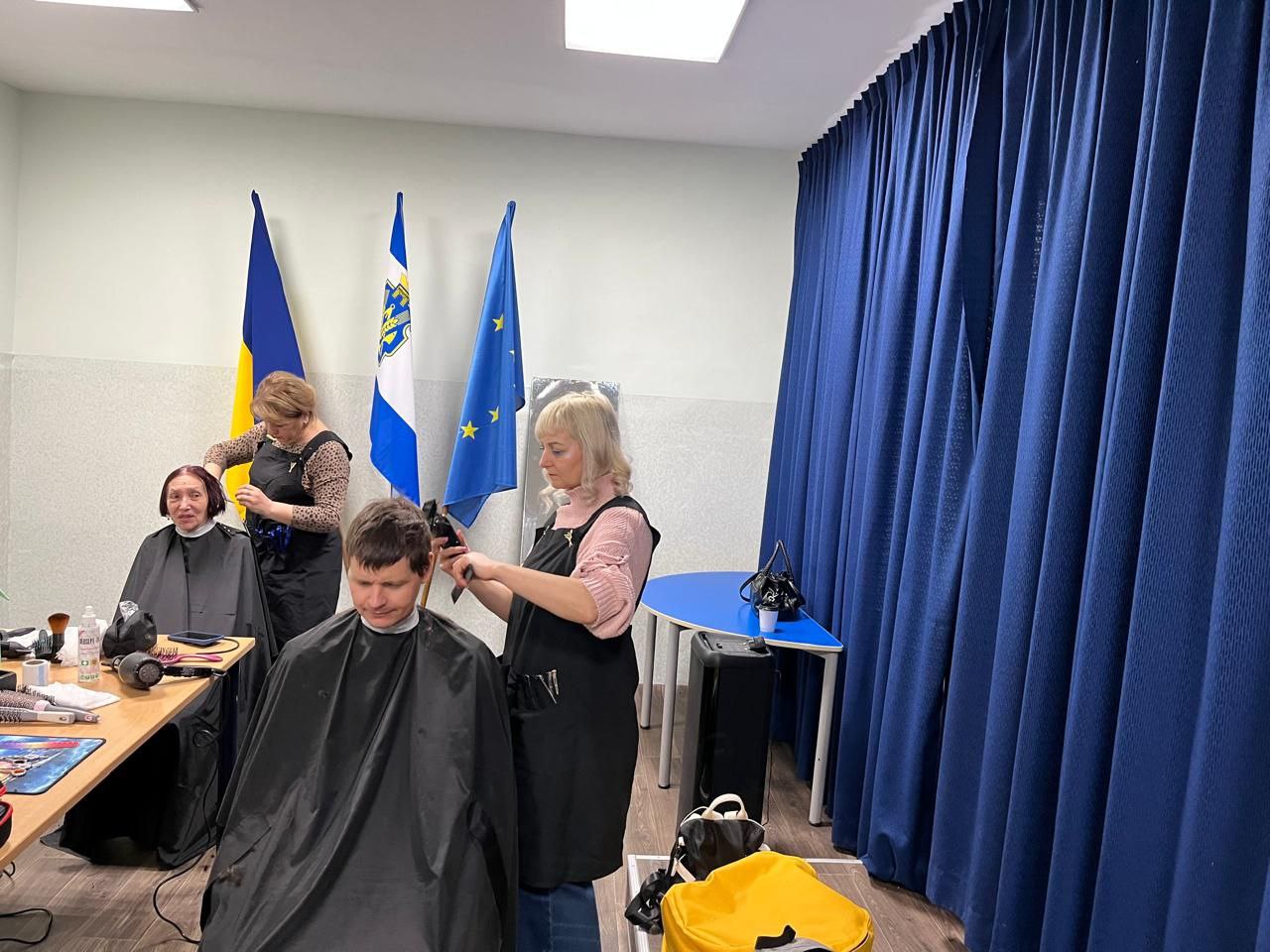 Двоє перукарів у чорних фартухах працюють над зачісками клієнтів. На задньому плані розташовані прапори України, Херсонської області та ЄС поруч із темно-синьою шторою.