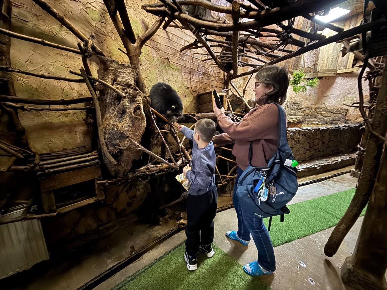 Дитина годує єнота у вольєрі, стилізованому під природне середовище з гілками та кам'яними стінами. Поруч стоїть жінка, яка знімає цей момент на смартфон.