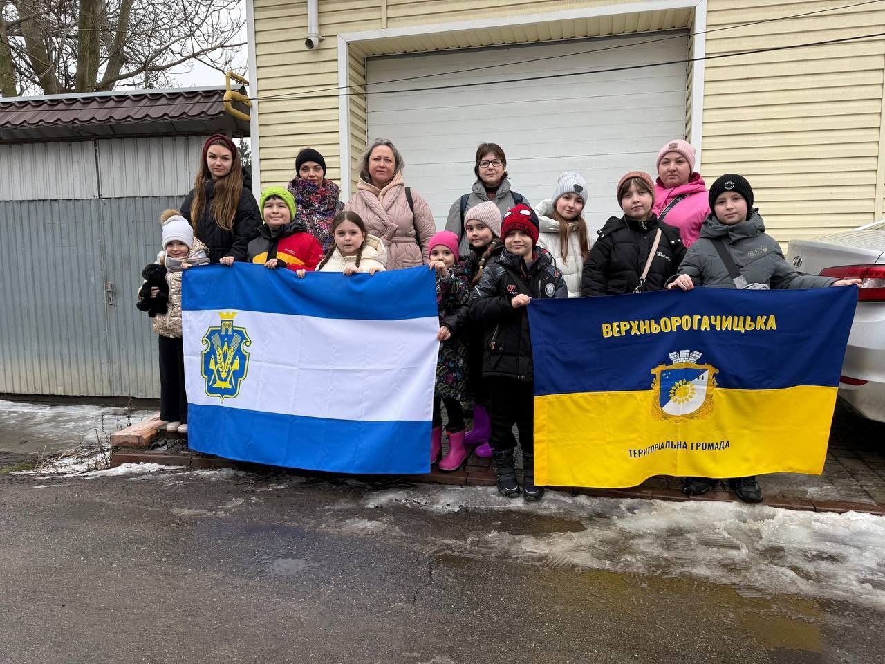 Група дорослих та дітей стоїть на вулиці поруч із будівлею, тримаючи два великі прапори. Один прапор — синьо-білий з гербом, інший — синьо-жовтий з написом «Верхньорогачицька територіальна громада» та символікою.