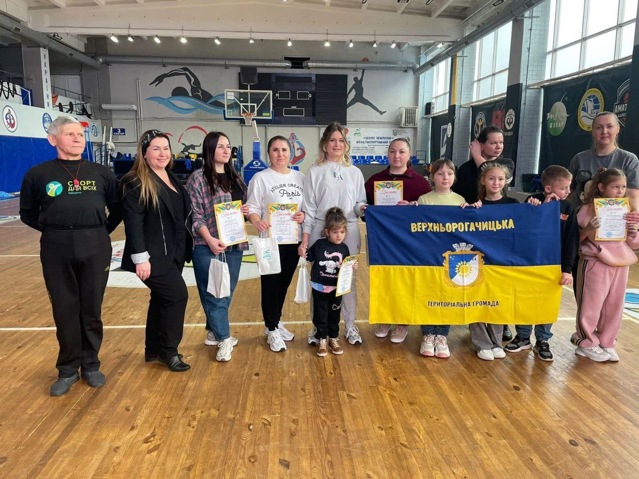 Група дорослих та дітей стоїть на дерев'яному покритті у спортивному залі. Деякі учасники тримають у руках грамоти та подарункові пакети. У центрі група тримає великий синьо-жовтий прапор Верхньорогачицької територіальної громади. На фоні видно баскетбольне кільце, спортивні плакати та вікна залу.